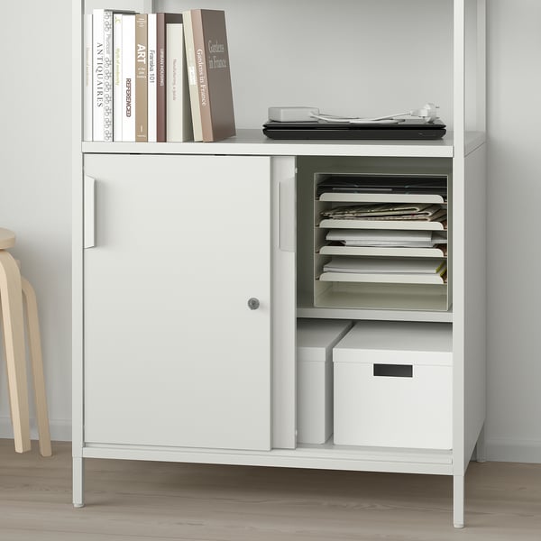 A white TROTTEN cabinet with adjustable shelves stores books, files, and a box, with sliding doors on one side for closed storage.
