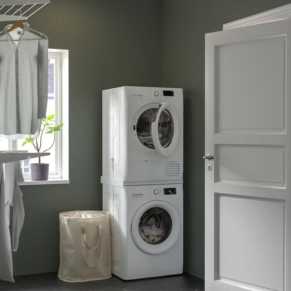 Compact white washing machine/dryer stack in laundry room with hamper and plant.