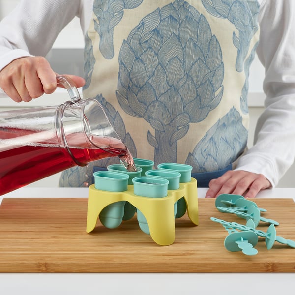 Person pouring red drink into green ice pop moulds on a wooden board.