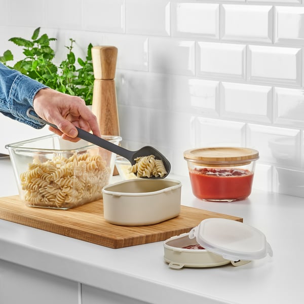 Person scooping noodles into a stackable lunch box on a wooden board. The lunch box has separate compartments for dry food.