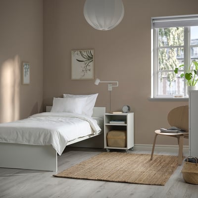 Minimalist bedroom with VIHALS bed, small table, and large light. Brown walls, wooden floor, chair, rug, and artwork.