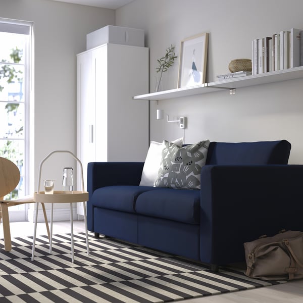 Navy blue VIMLE sofa bed by large window, on striped rug, with side table and shelf.