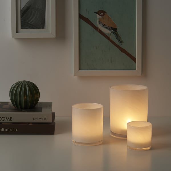 Three VINDSTILLA candles in frosted glass on table with books, smallest in front, translucent glass.