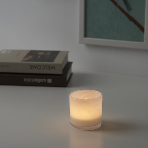 Lit candle in glass holder on table, warm glow, with books and framed picture.