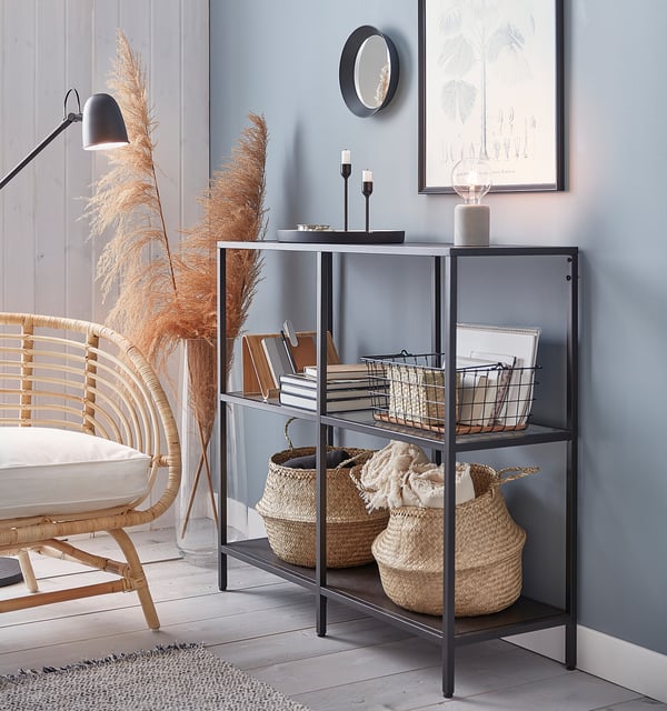 Black metal shelf with glass, baskets and books. Soft blue wall, white art and wicker chair.