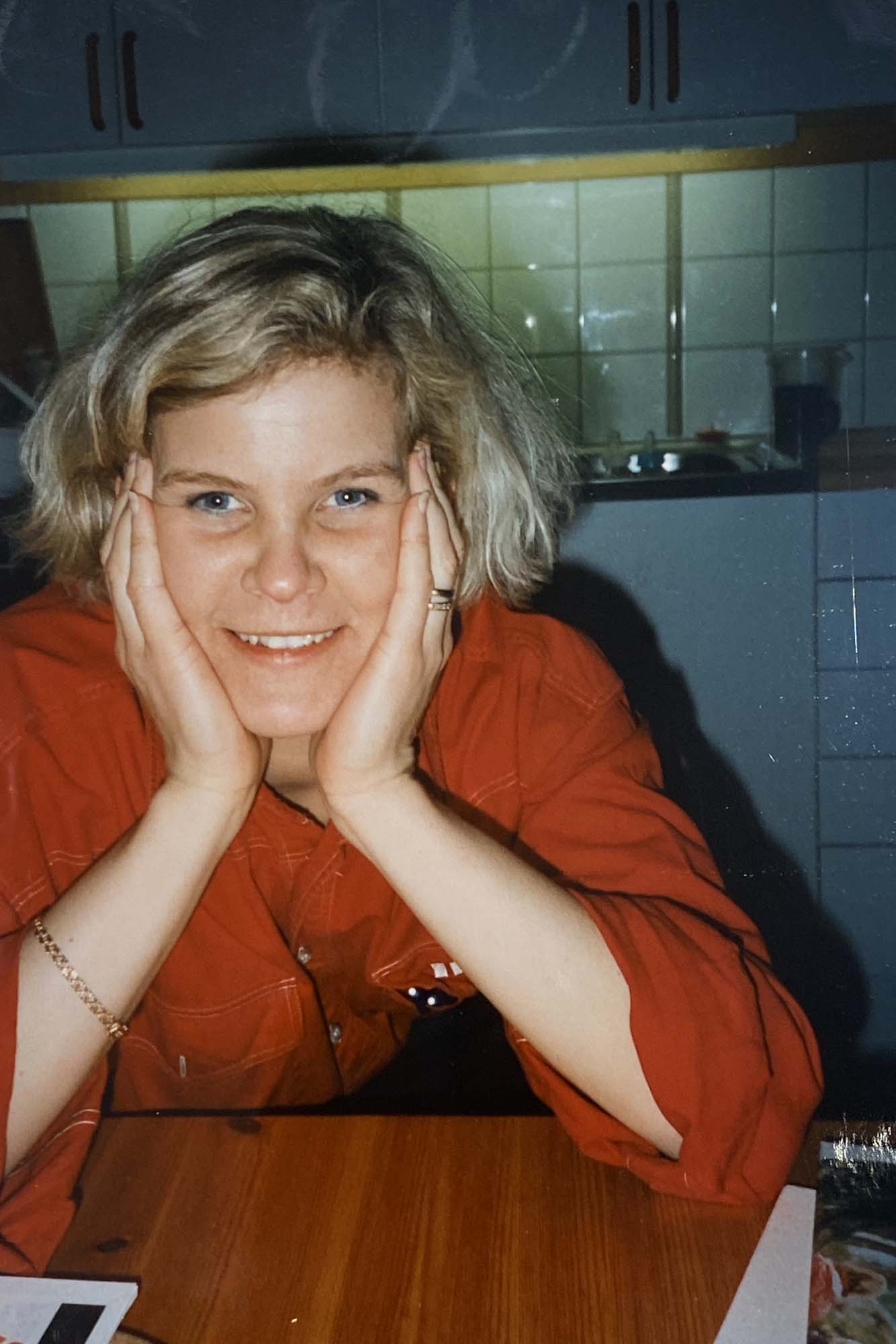 A vintage image of Fillipa sitting in her kitchen wearing a red IKEA uniform. She sits with her hands in her face, smiling.