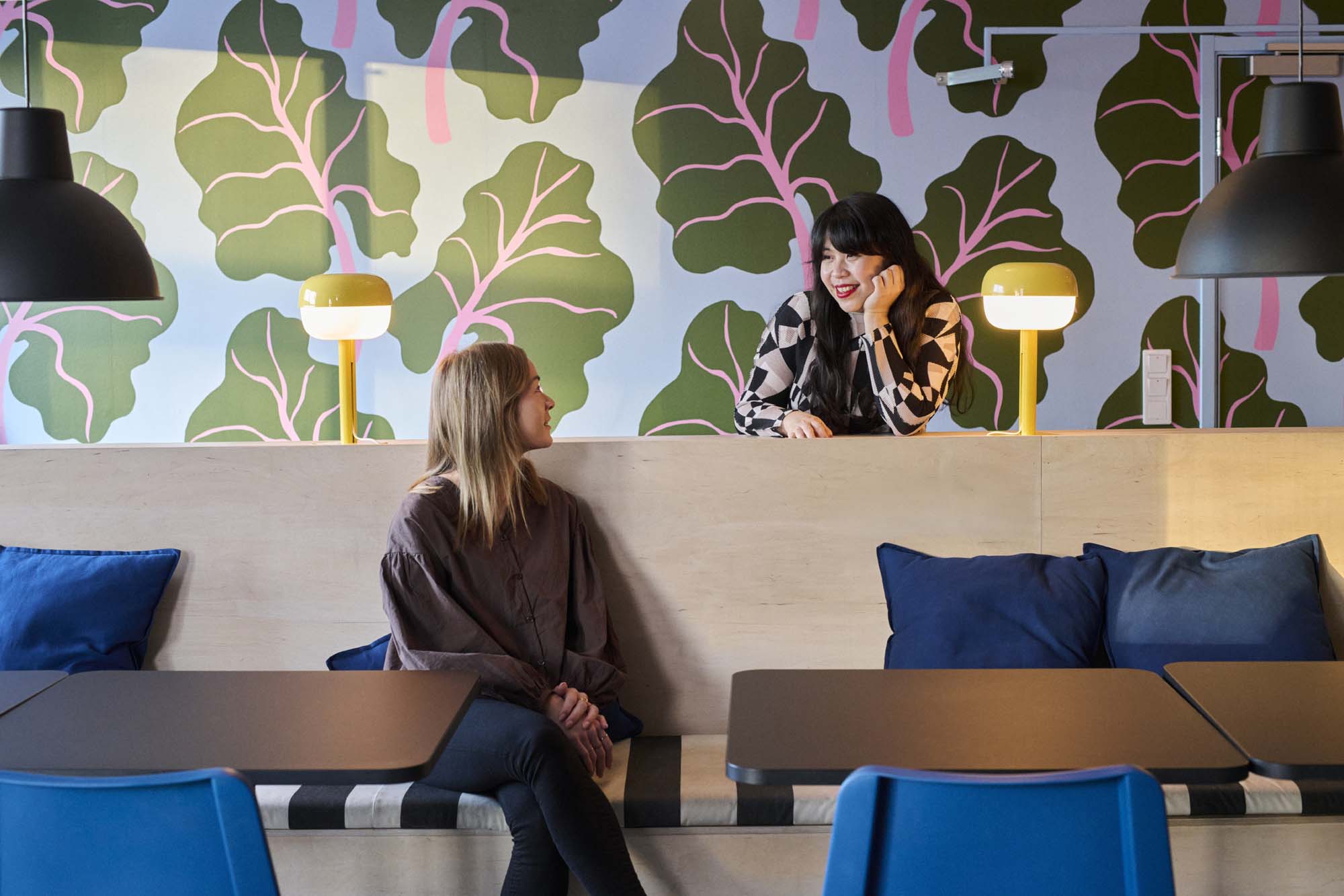 A person leans on the back of a wooden bench in a brightly coloured work environment. They are talking to a person sitting on the wooden bench in front.