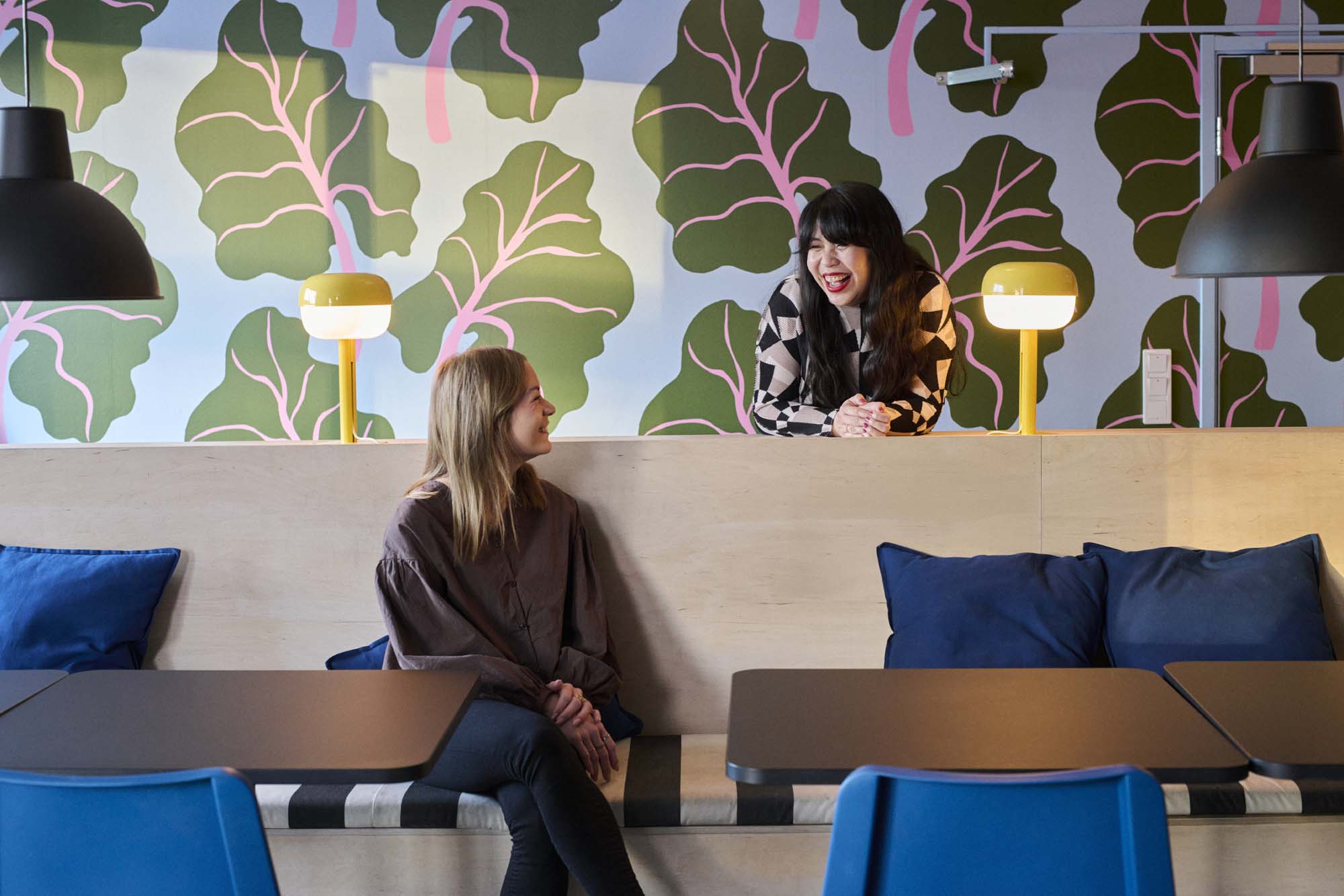 Two people are speaking. One leans on a wooden counter behind the other who sits and looks over their shoulder to speak.