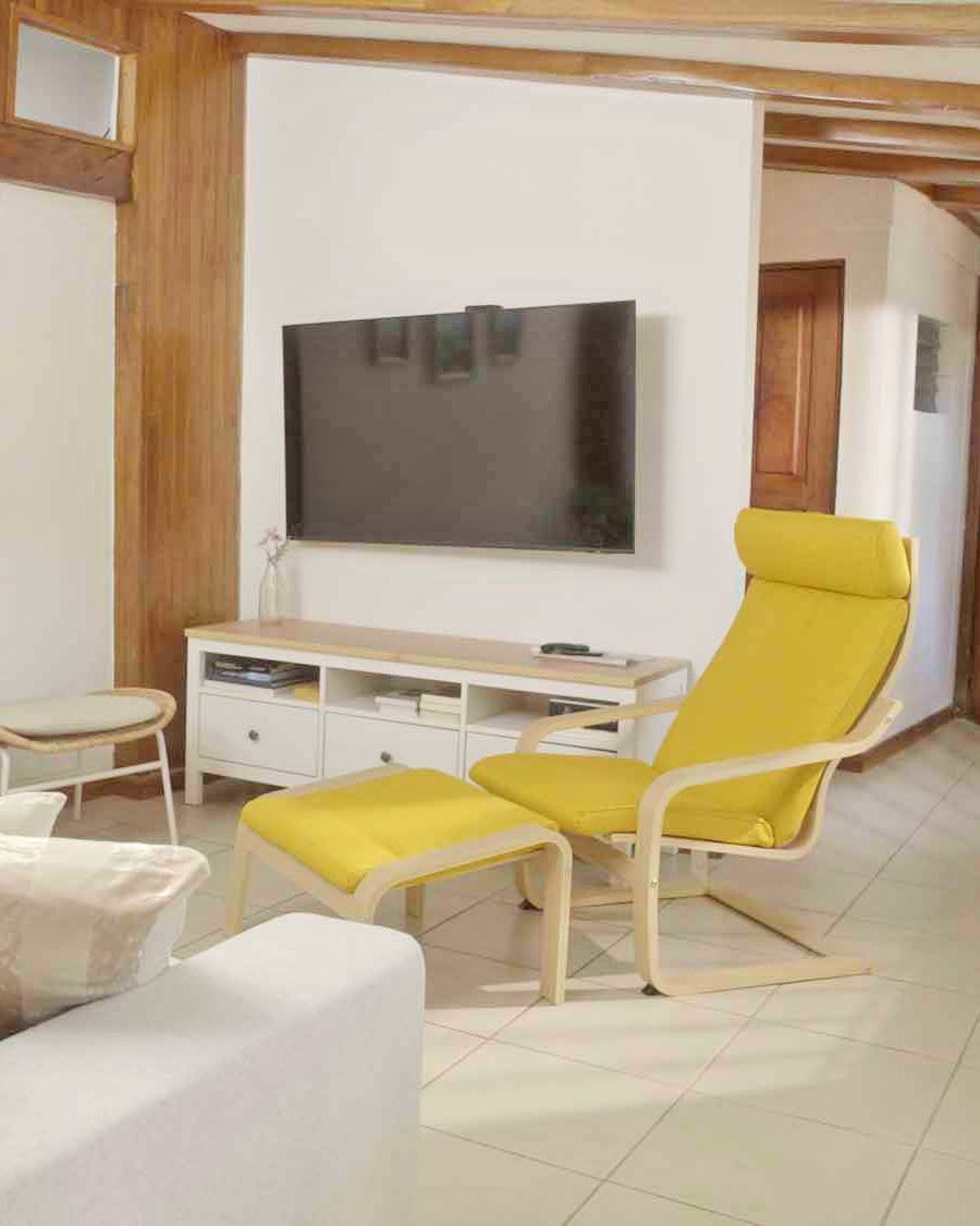 A living room with a yellow chair and footstool in front of a wall-mounted TV.