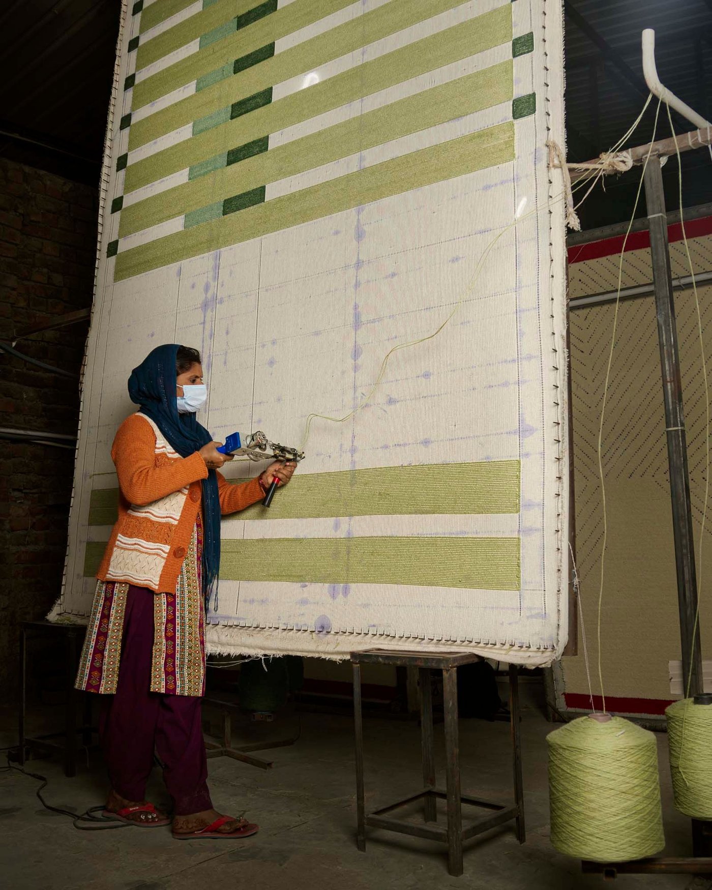 A person wearing a face mask stands in front of a giant rug canvas. They are holding a tufting gun against the canvas.