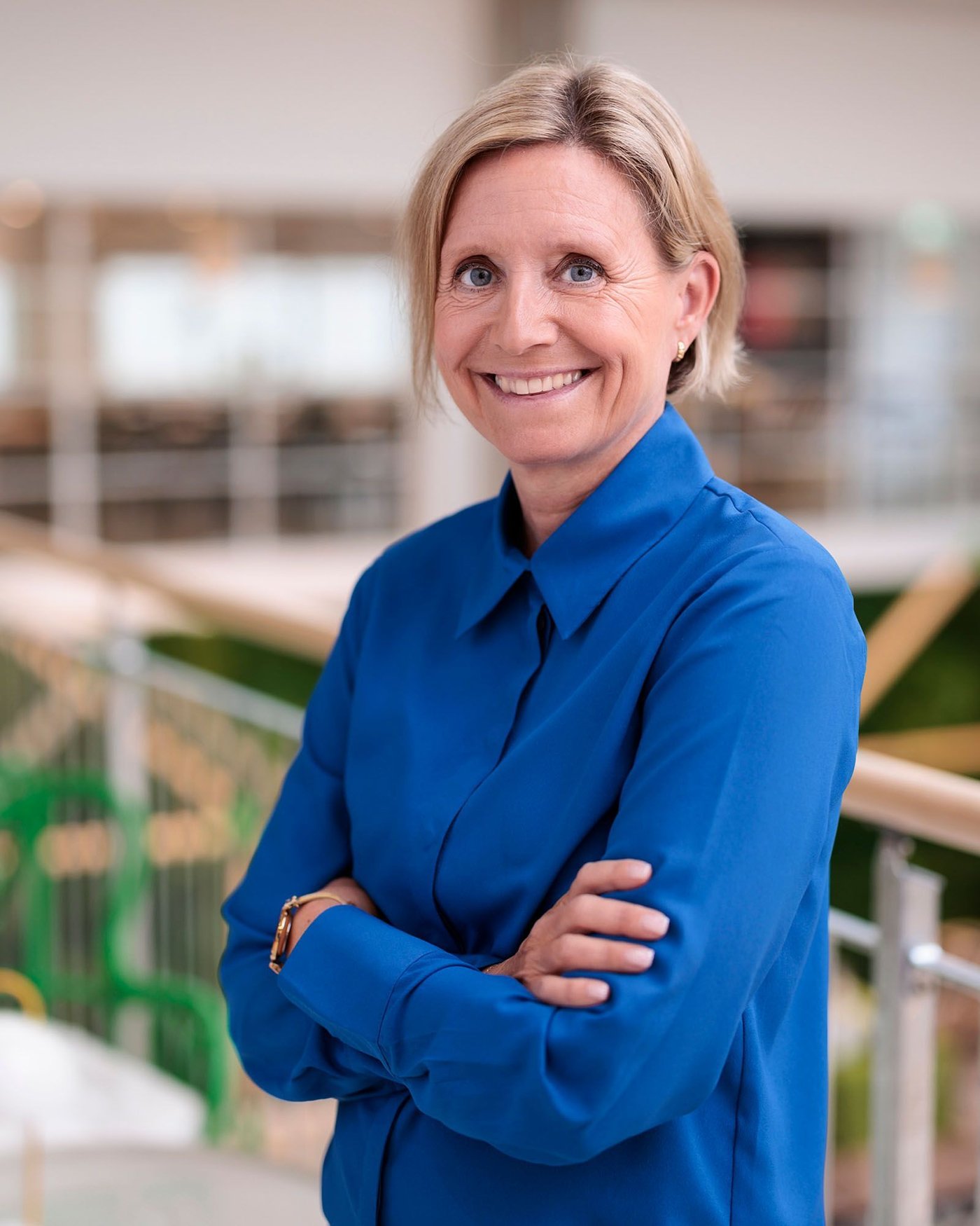 Portrait of Åsa Skogström Feldt, wearing a cobalt blue shirt and standing with arms folded in a bright office environment.