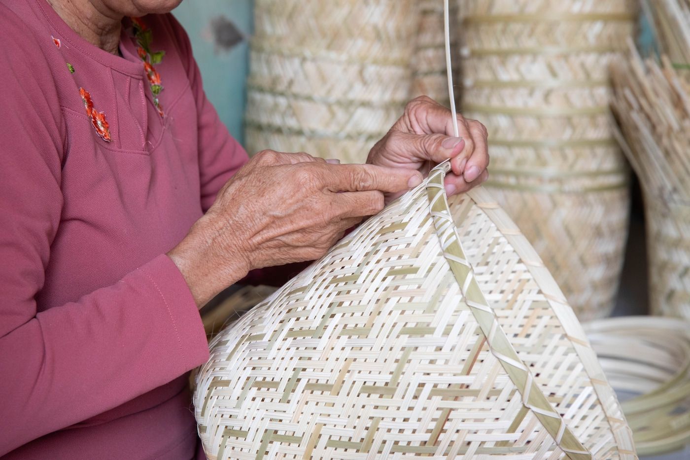 A person weaves a rattan basket next to a stack of similar baskets.
