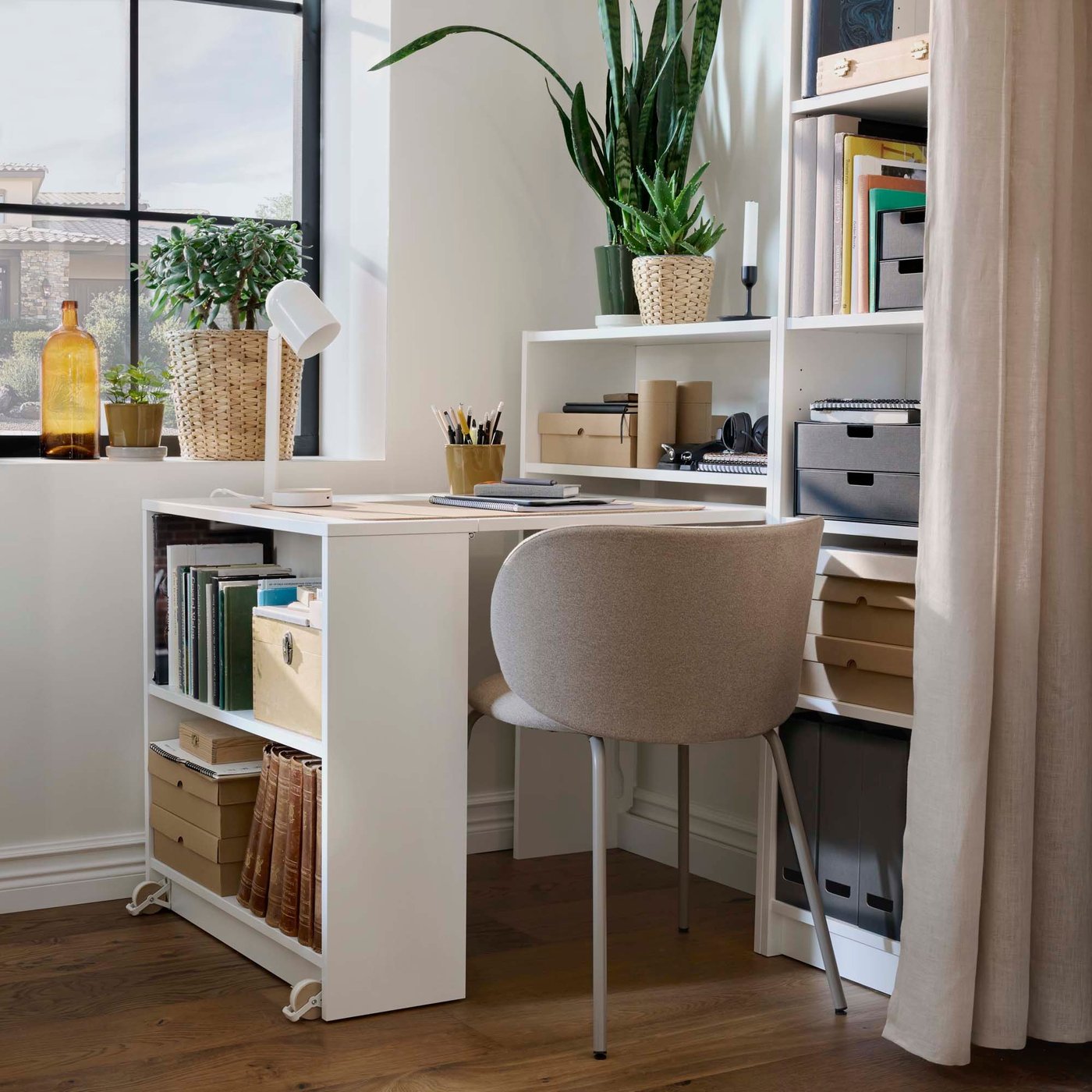 A home office with a white desk connected to a bookcase, shelves filled with books and storage boxes, and a potted plant in front of a window.