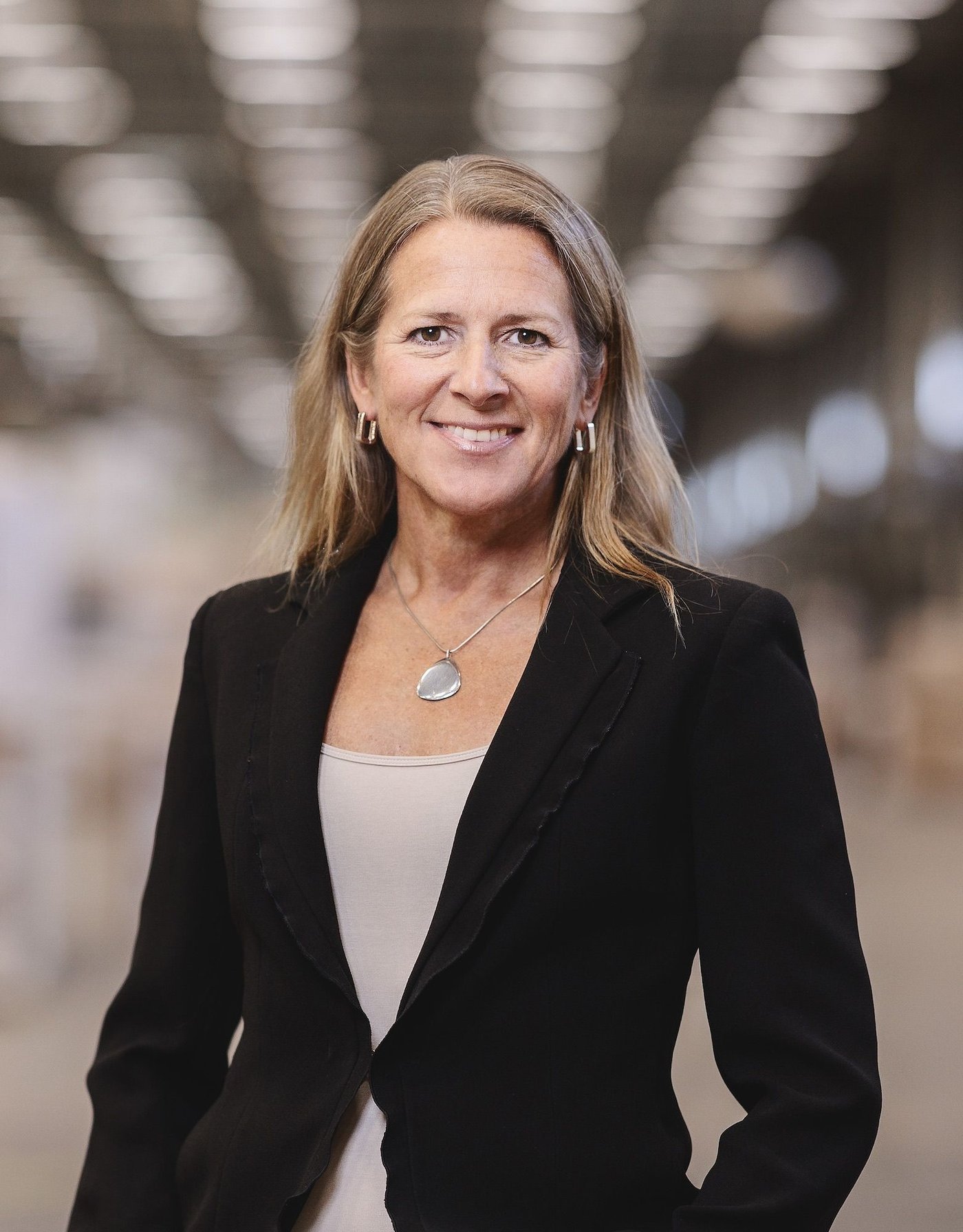 Portrait of Christina Niemelä Ström, who is wearing a black blazer with a blurred industrial space in the background.