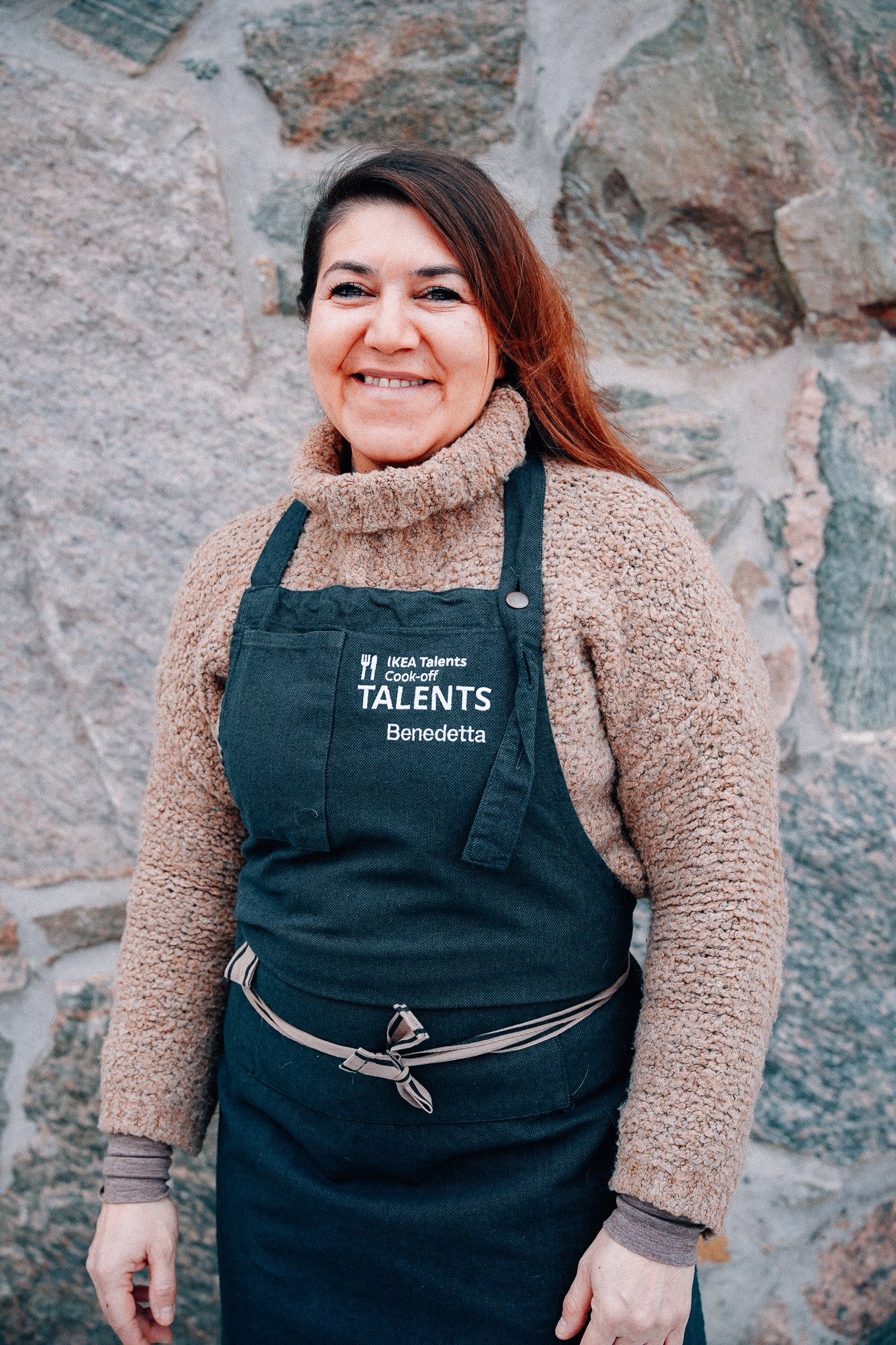 A person wearing a dark apron over a sweater stands in front of a textured stone wall. The apron features embroidered text, that reads "IKEA Talents Cook-off.