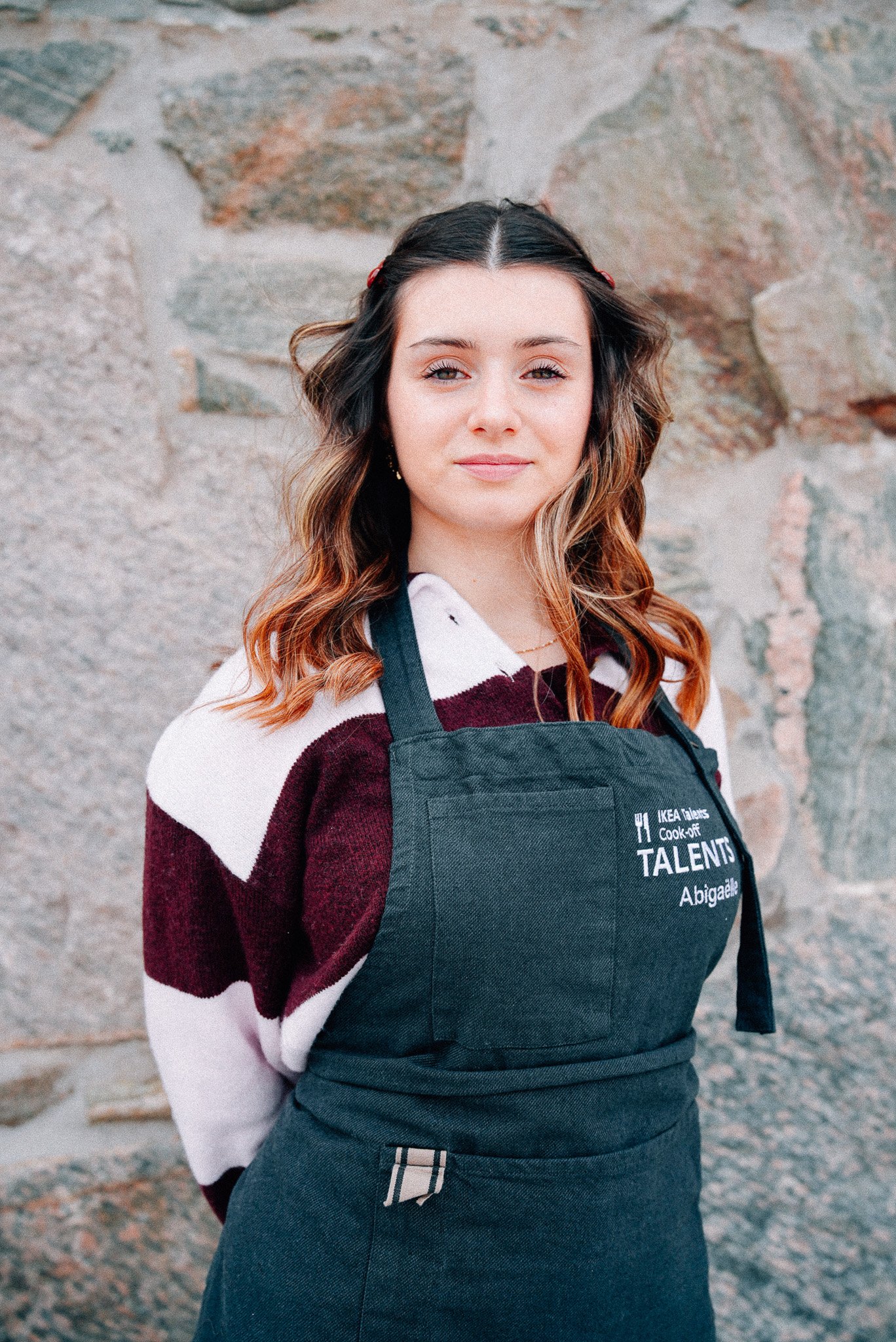 A person wearing a dark apron over a striped sweater stands in front of a textured stone wall. The apron features embroidered text, that reads "IKEA Talents Cook-off.