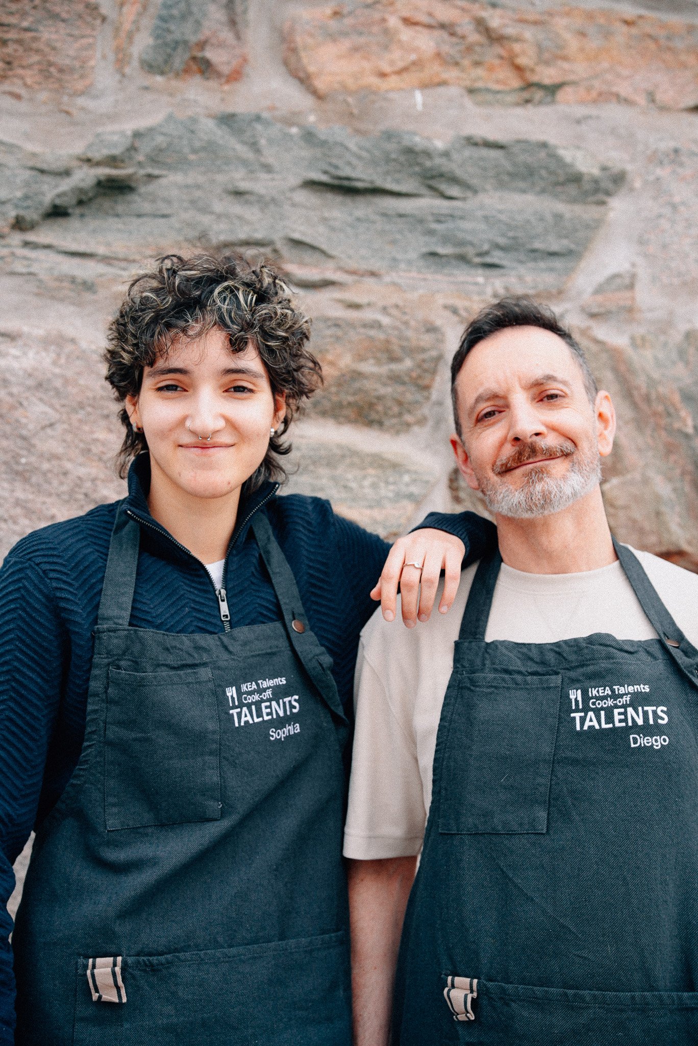 Two people wearing dark aprons over sweaters, standing in front of a textured stone wall. The apron features embroidered text, that reads "IKEA Talents Cook-off.