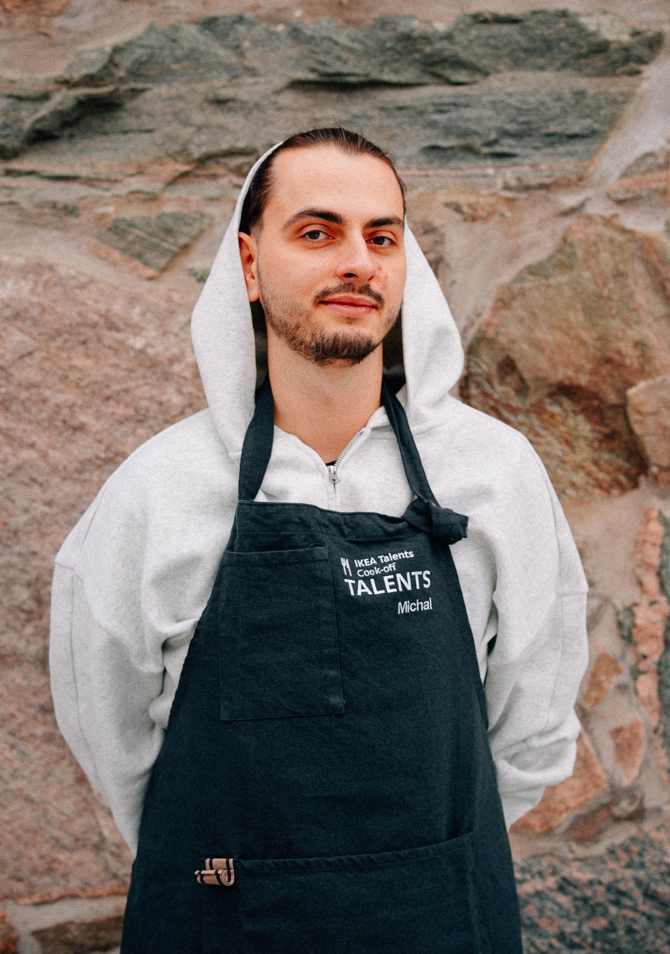 A person wearing a dark apron over a sweater stands in front of a textured stone wall. The apron features embroidered text, that reads "IKEA Talents Cook-off.