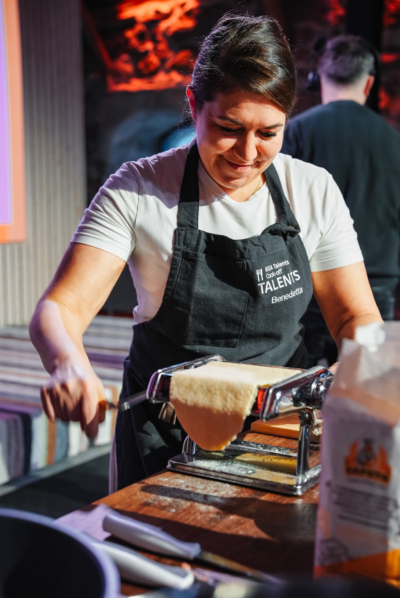 A person wearing an apron uses a pasta machine to roll out dough in a kitchen setting. The scene includes various cooking ingredients and a warm ambiance with soft, diffused lighting.