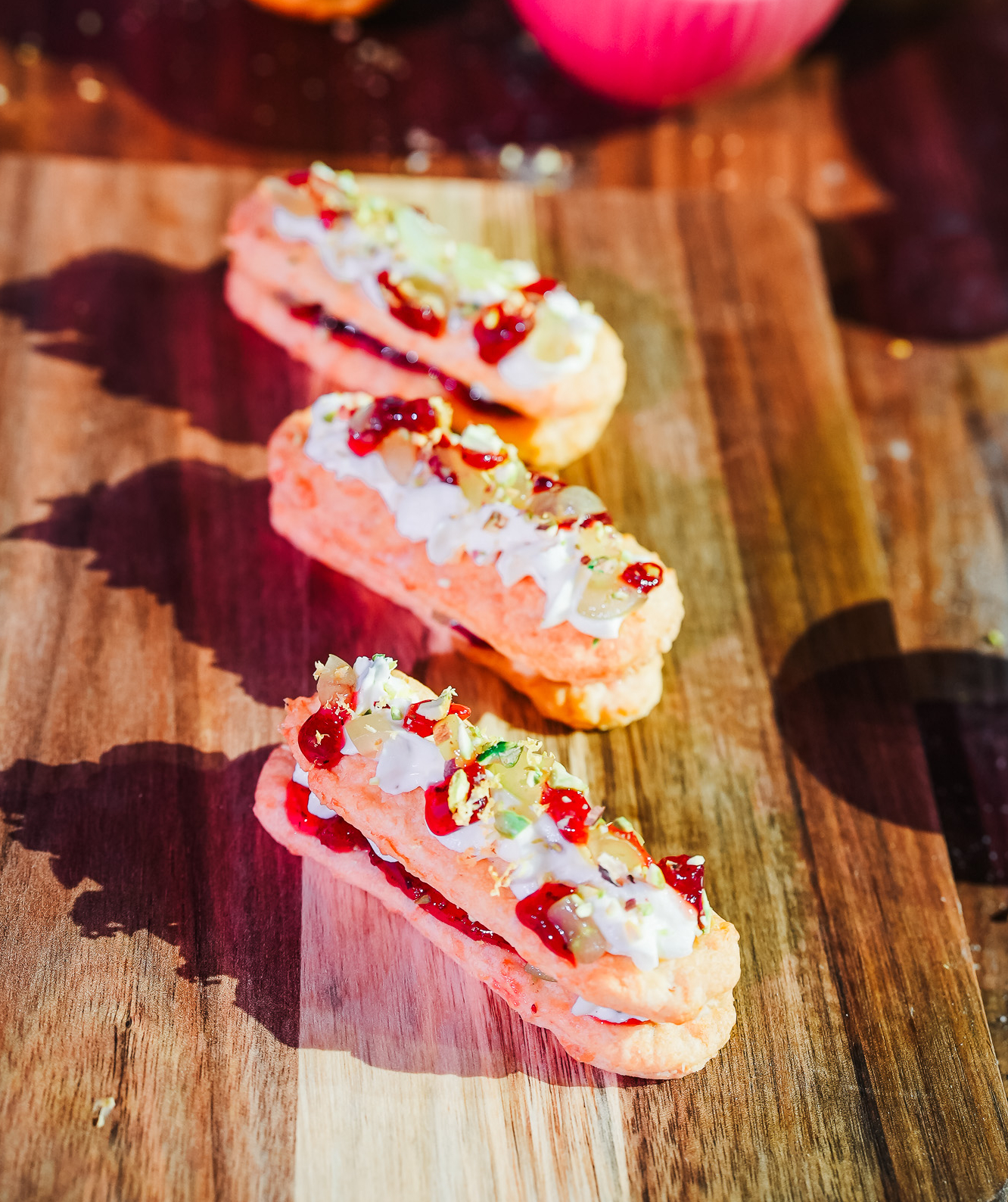 Three pastries topped with cream and red fruit pieces are arranged on a wooden surface, creating an appetising visual with shadows cast by sunlight.
