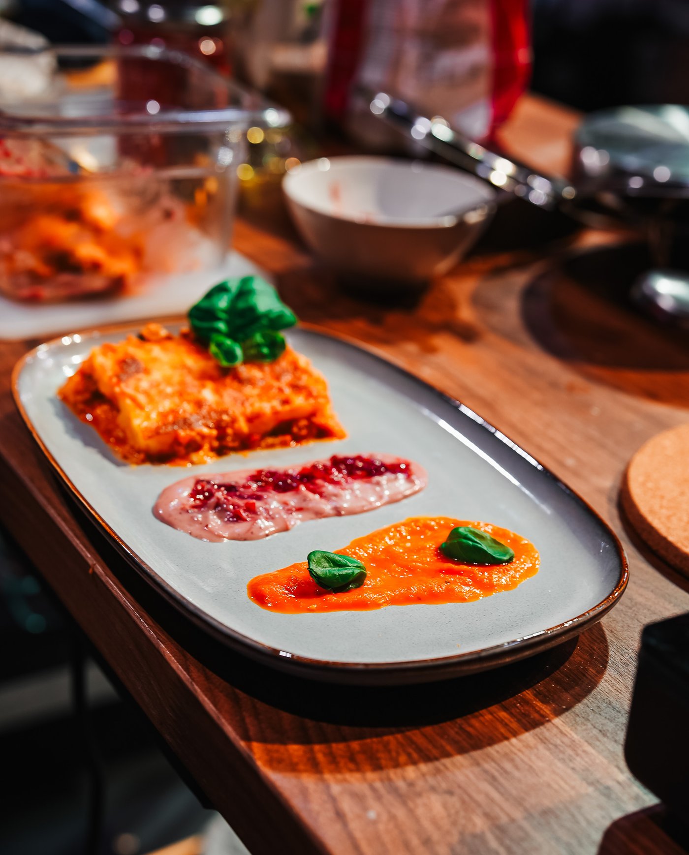 An oval plate on a wooden table features a piece of lasagna with a savory portion, complemented by colourful sauces and garnished with herbs. Surrounding utensils and ingredients add to the culinary presentation.