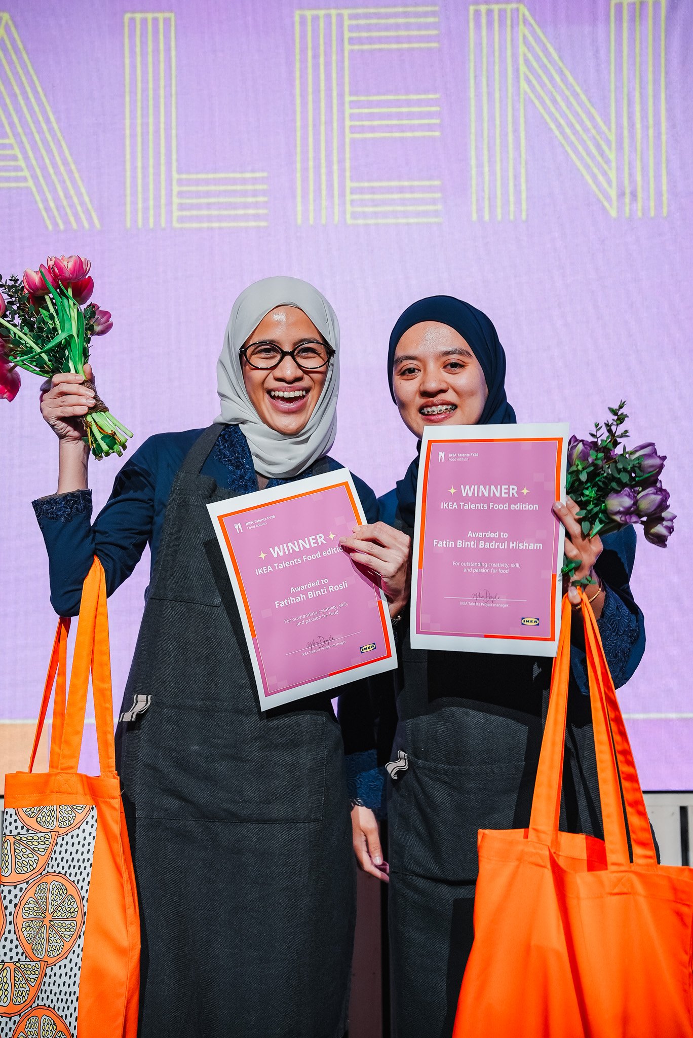 Two people hold certificates and bouquets, standing in front of a purple backdrop. They carry bright orange bags, adding a vibrant contrast to their dark aprons and festive setting.