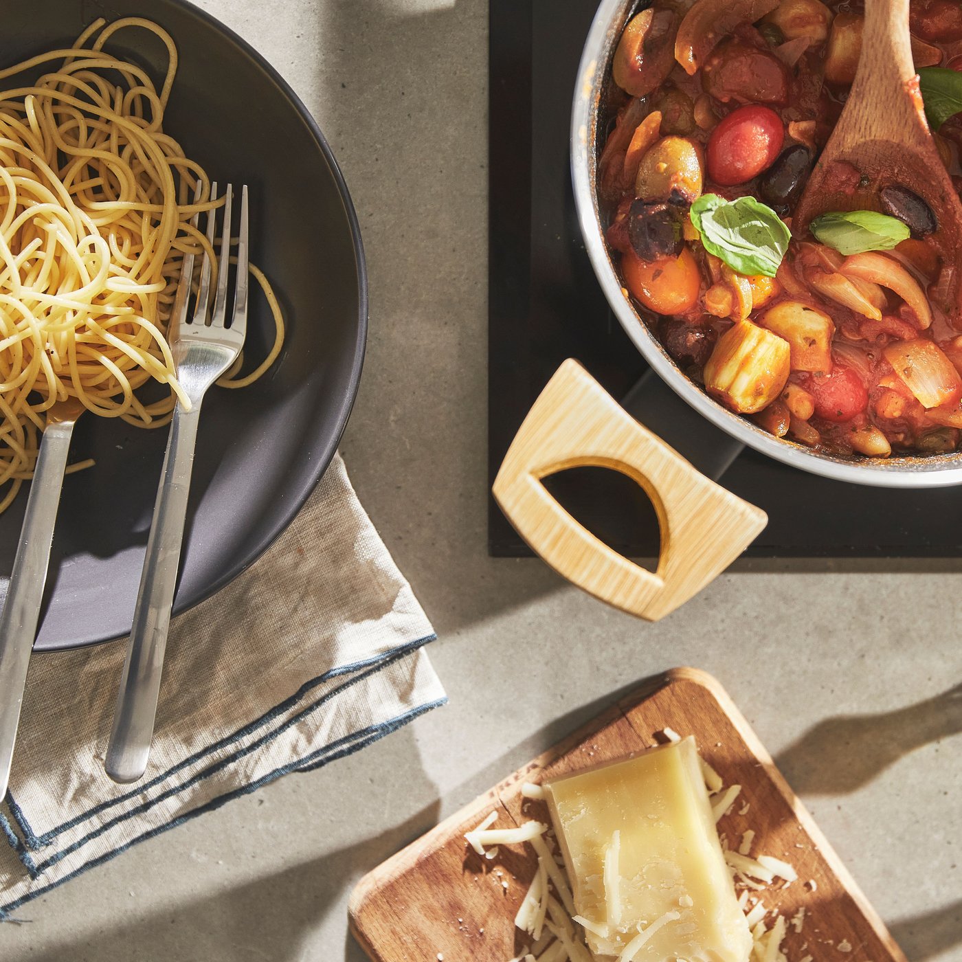 One pan filled with plain cooked spaghetti. Next to it a pan with the pasta sauce consisting of different vegetables. Next to that a wooden chop board with some parmesan cheese.