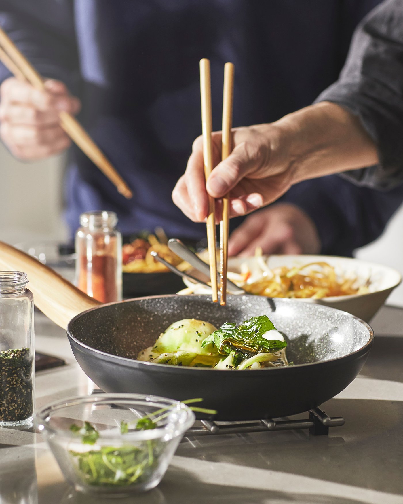 The HUSKNUT frying pan filled with some green vegetables. Someone holding two chopsticks right above the pan. In the background some other dishes and in front of the pan a small glass bowl filled with some small chopped greens.