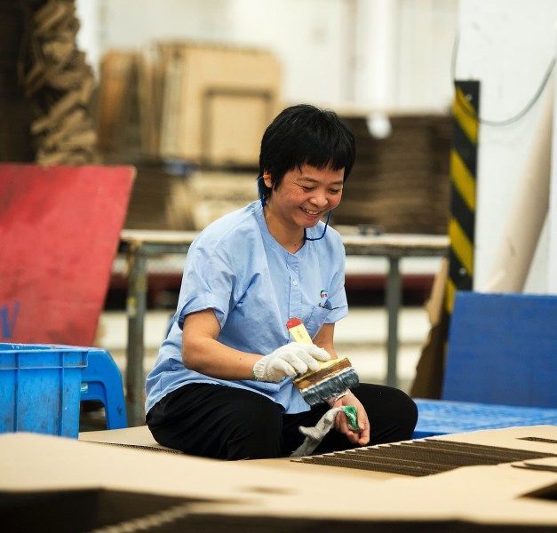 In a factory environment, a person is sitting with a brush in one hand hand and a glove in the other hand.