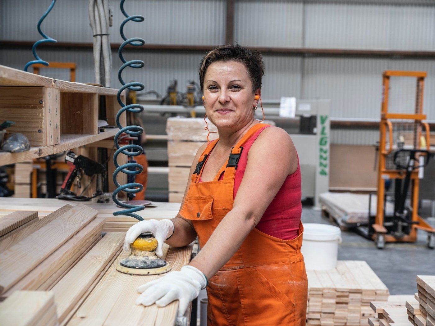 A person in orange workwear standing in an industry environment sanding a plank.