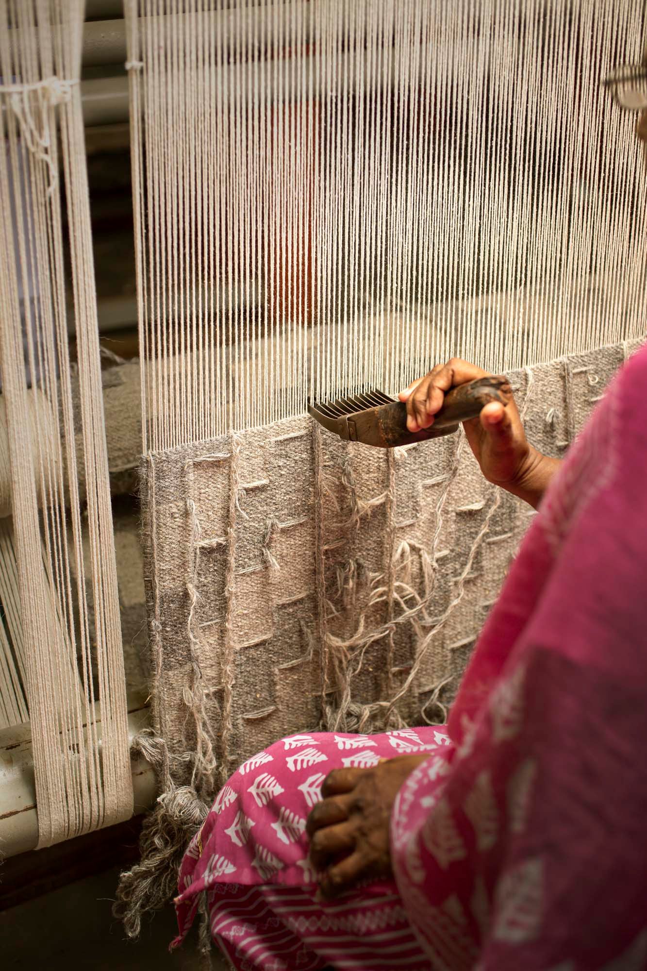 A person sits at a vertical loom, using a panja tool to push down the threads.