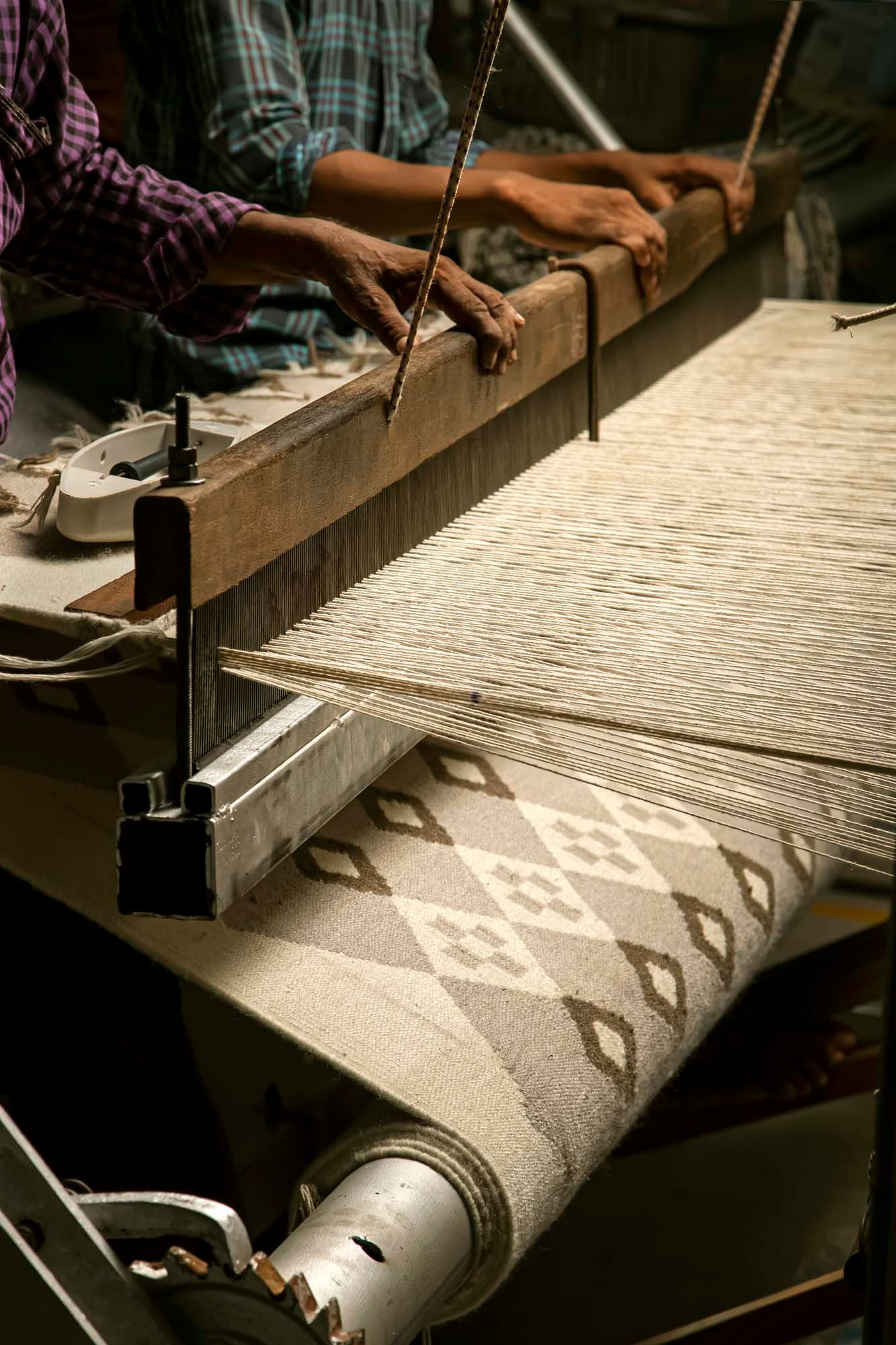 Two pairs of hands work a loom, creating a diamond patterned rug.