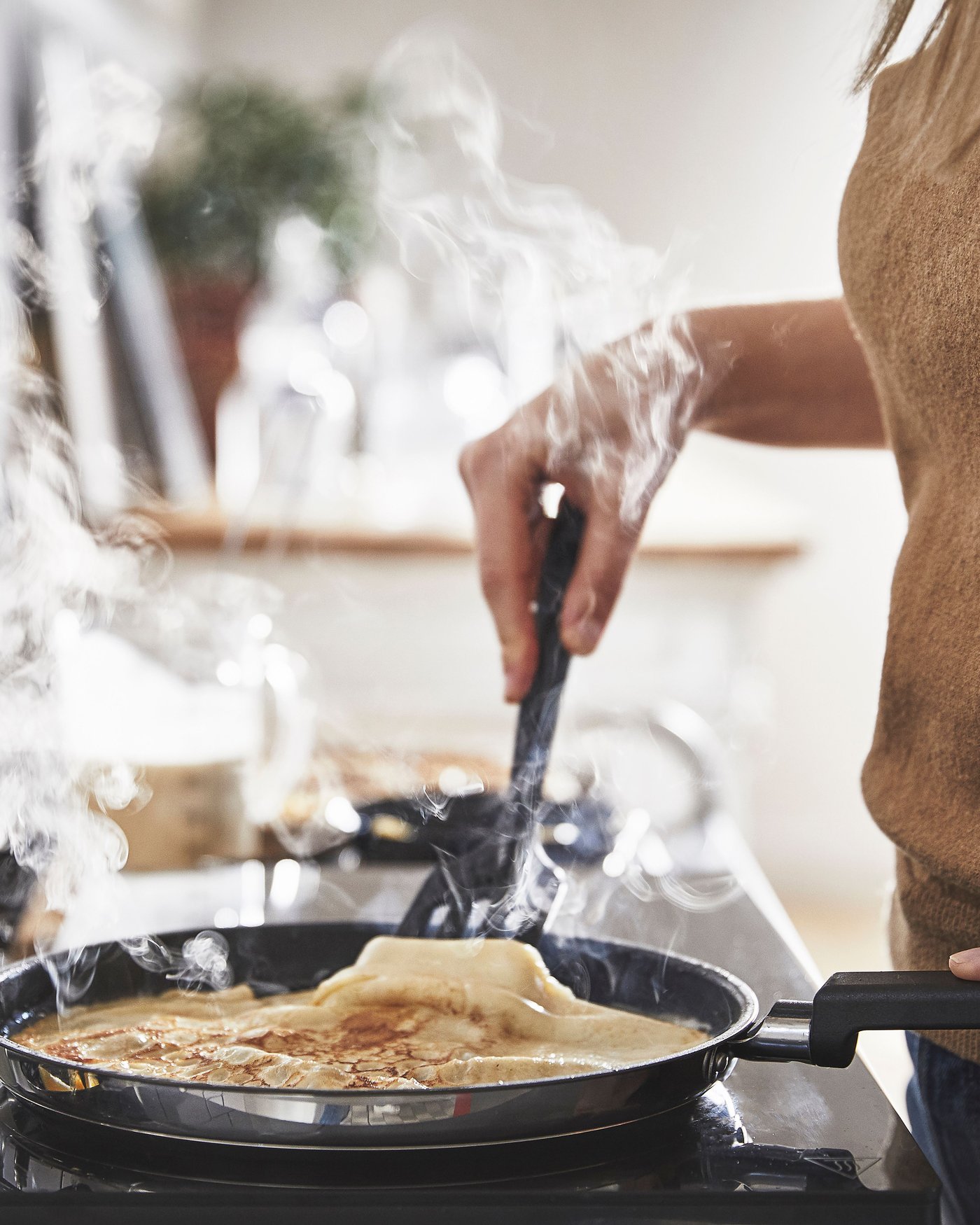 The MIDDAGSMAT pan on the stove. In the pan there's a pancake. Someone is holding the pan and has a cooking utility underneath the pancake, holding the pancake up a little.