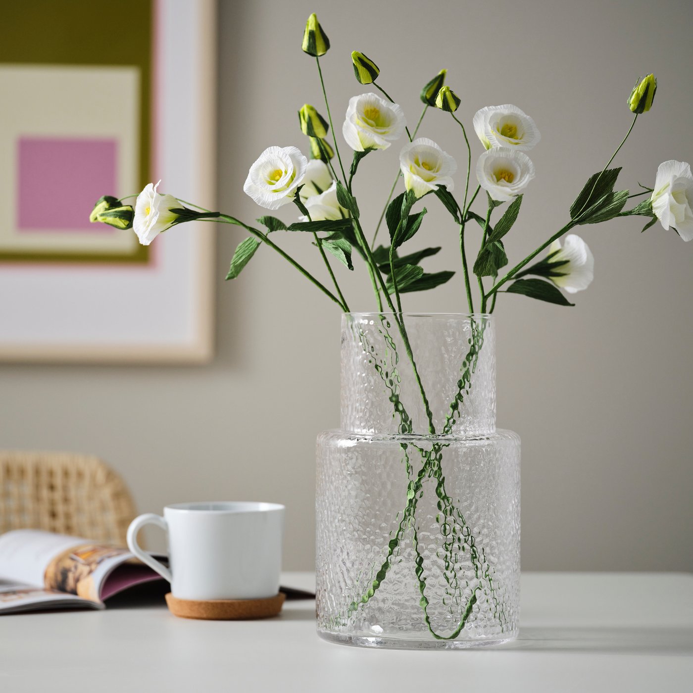 A dining room with a clear vase filled with white GULDSPETT artificial flowers, placed on a table with a painting in the background.