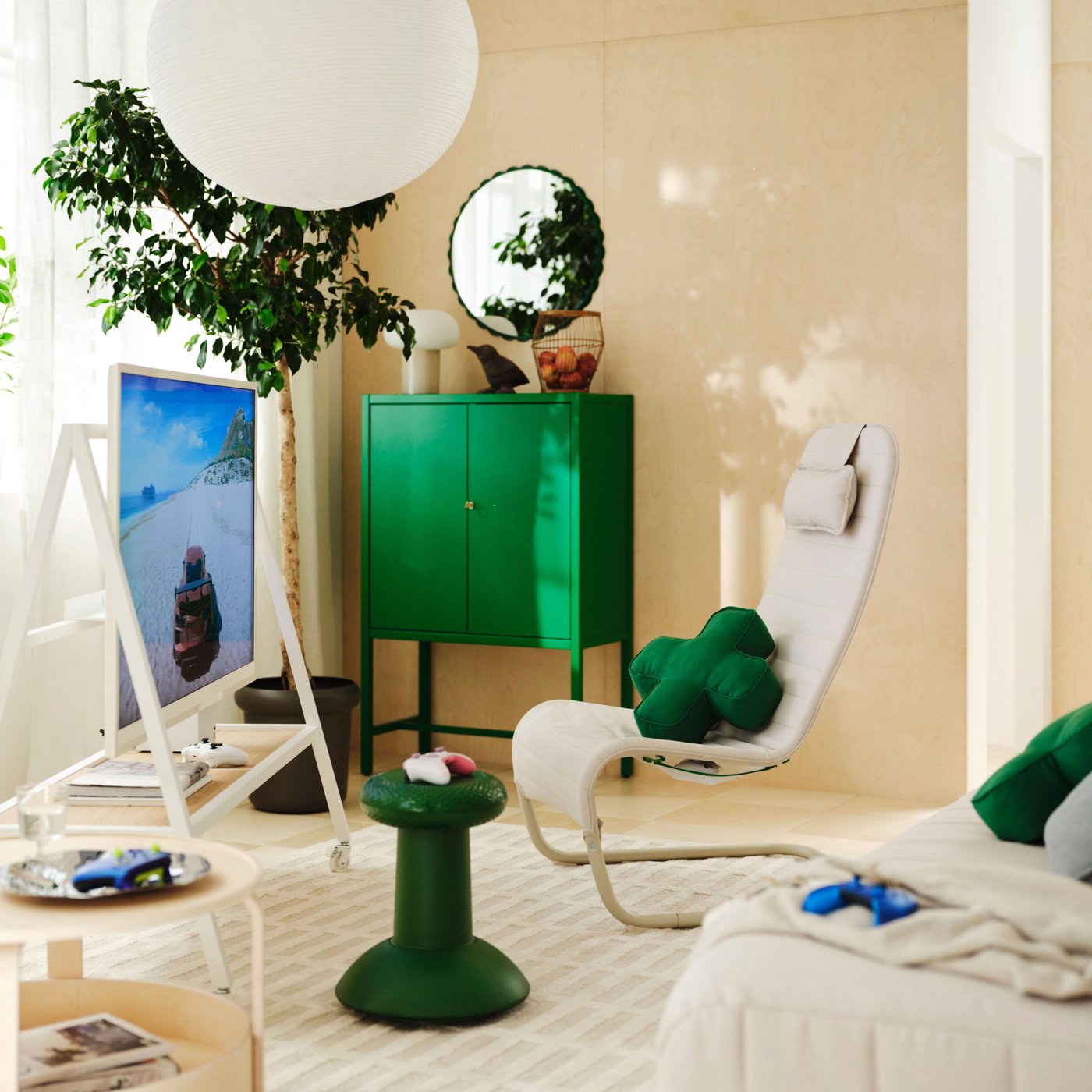 A bright, sunlit living room with a white gaming lounge chair, green storage cabinet and side table, large paper pendant lamp, indoor plants, and a TV displaying a beach scene.
