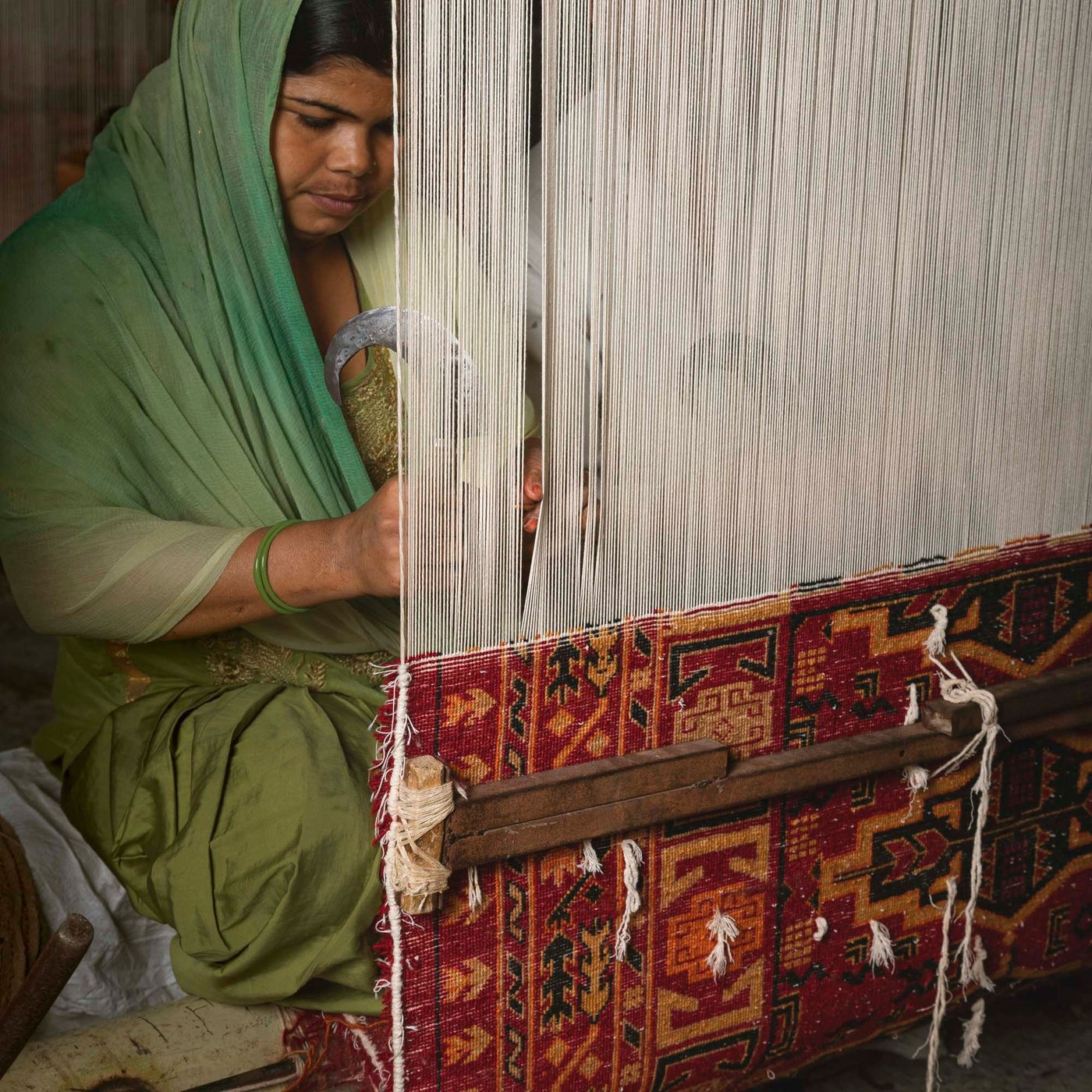 A person stands at a vertical loom, working on a very decorative red rug.