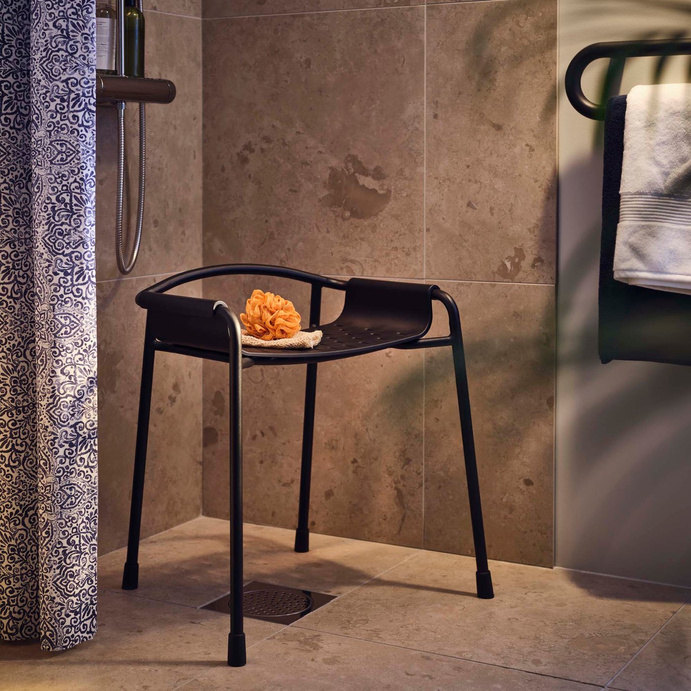 A black BÄSINGEN shower chair in the corner of shower with beige tiles.