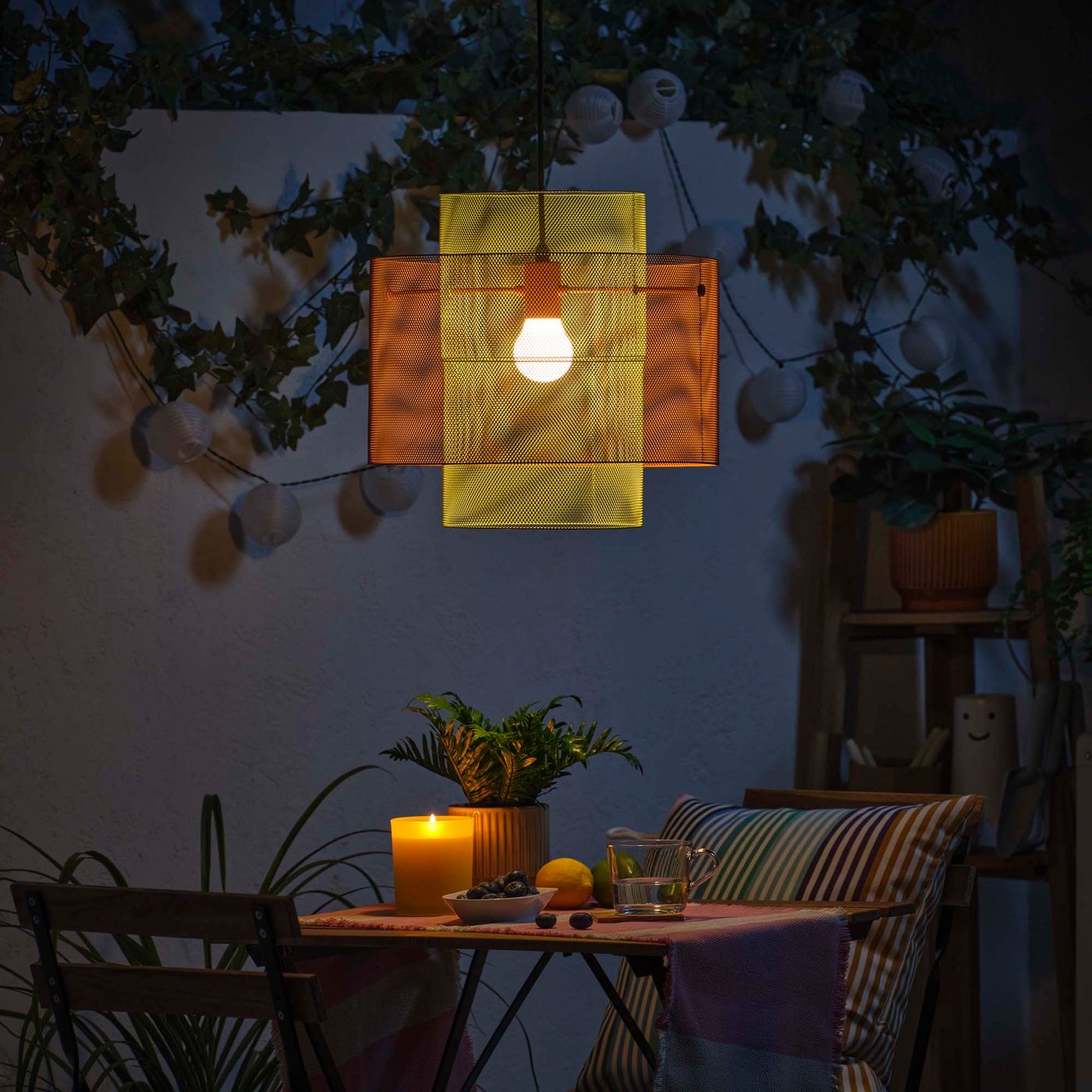 A glowing yellow and orange mesh pendant ceiling lamp hangs over an outdoor table in the dark. 