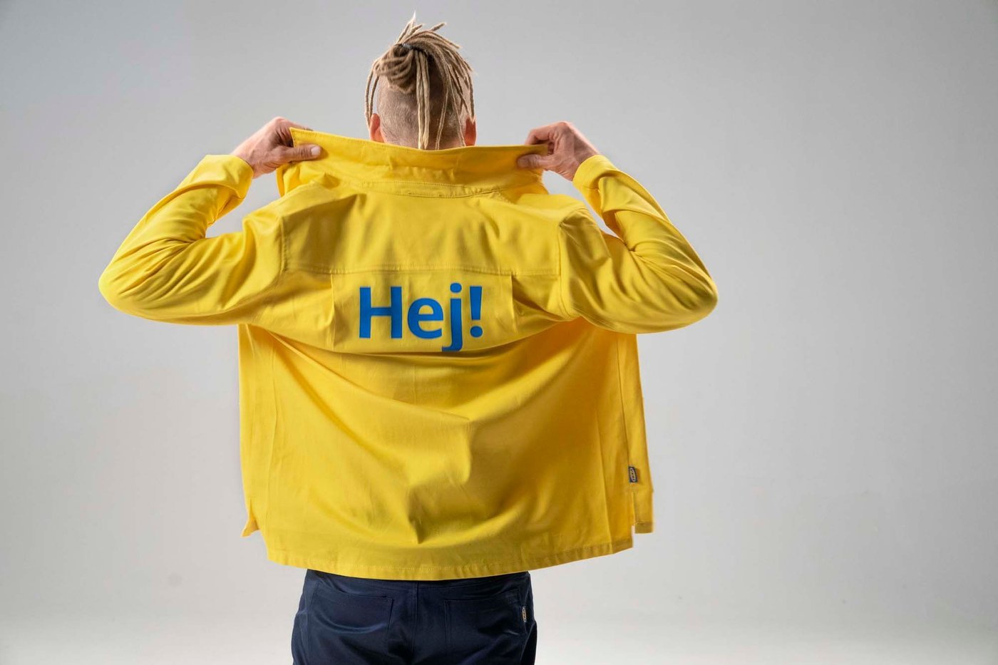 The back of a person raising the collar of their yellow jacket with "Hej!" written in blue across the shoulders.