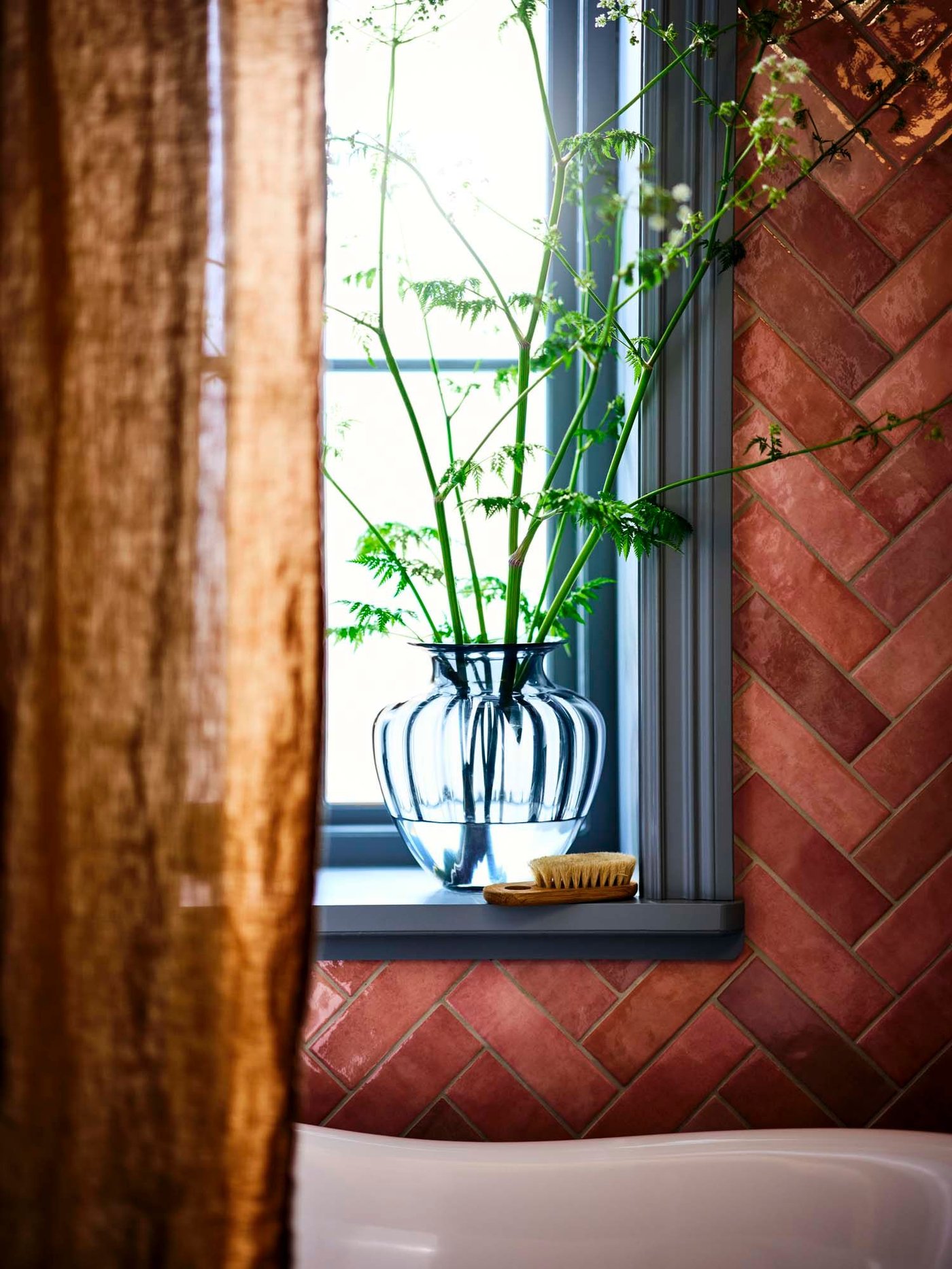 A sheer brown curtain is pulled back, revealing a large blue glass vase filled with green stems on a window ledge.
