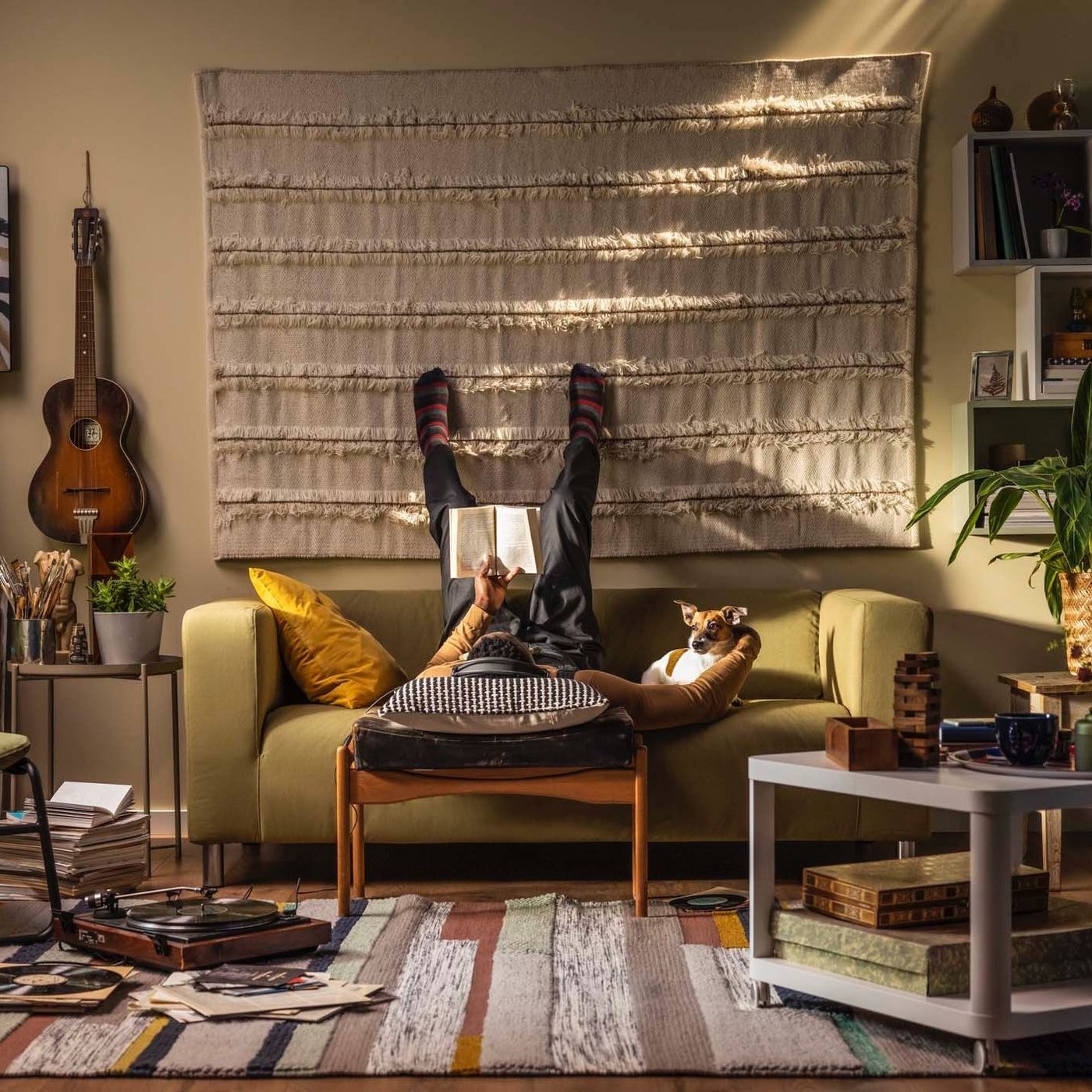 A living area with a person and a dog on a sofa. The person is lying down, reading, with their legs up the wall behind the sofa.