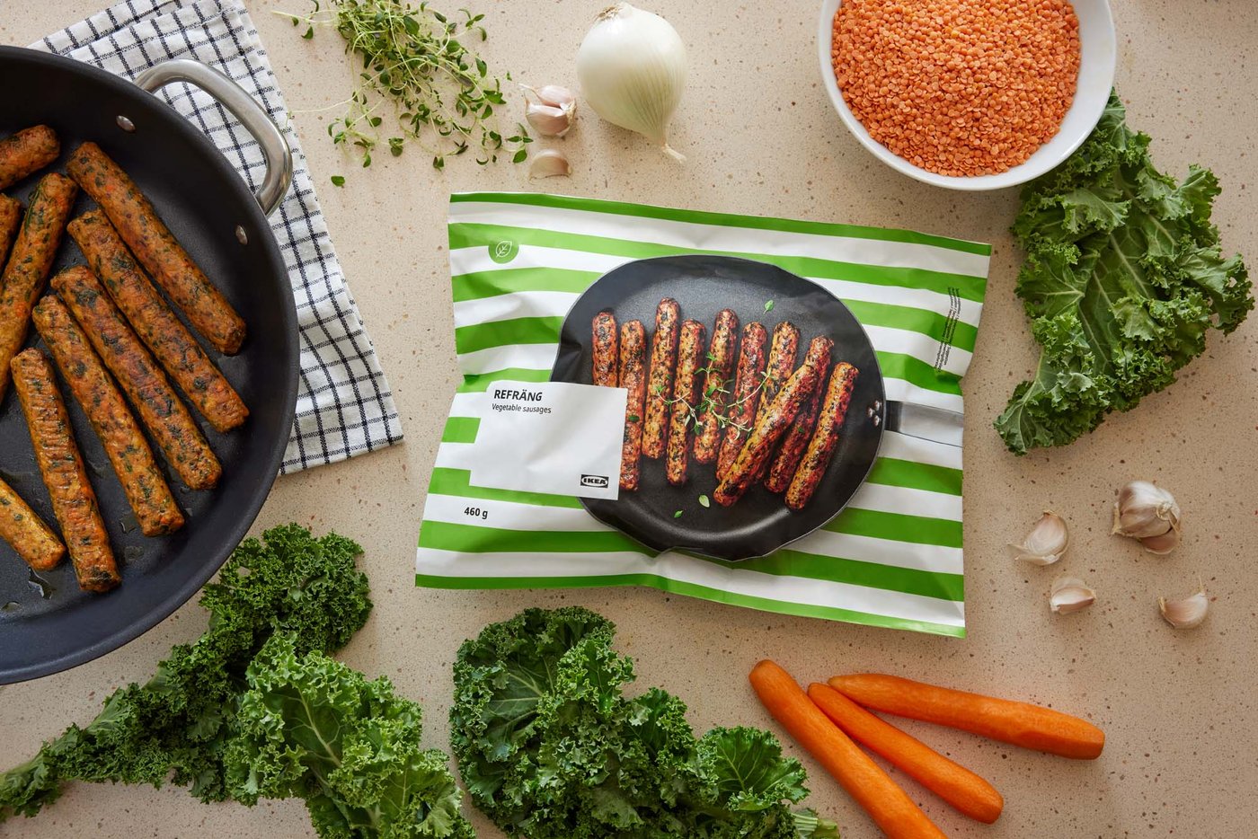 A pack of REFRÄNG vegetable sausages on a surface, surrounded by ingredients and a pan of sausages.