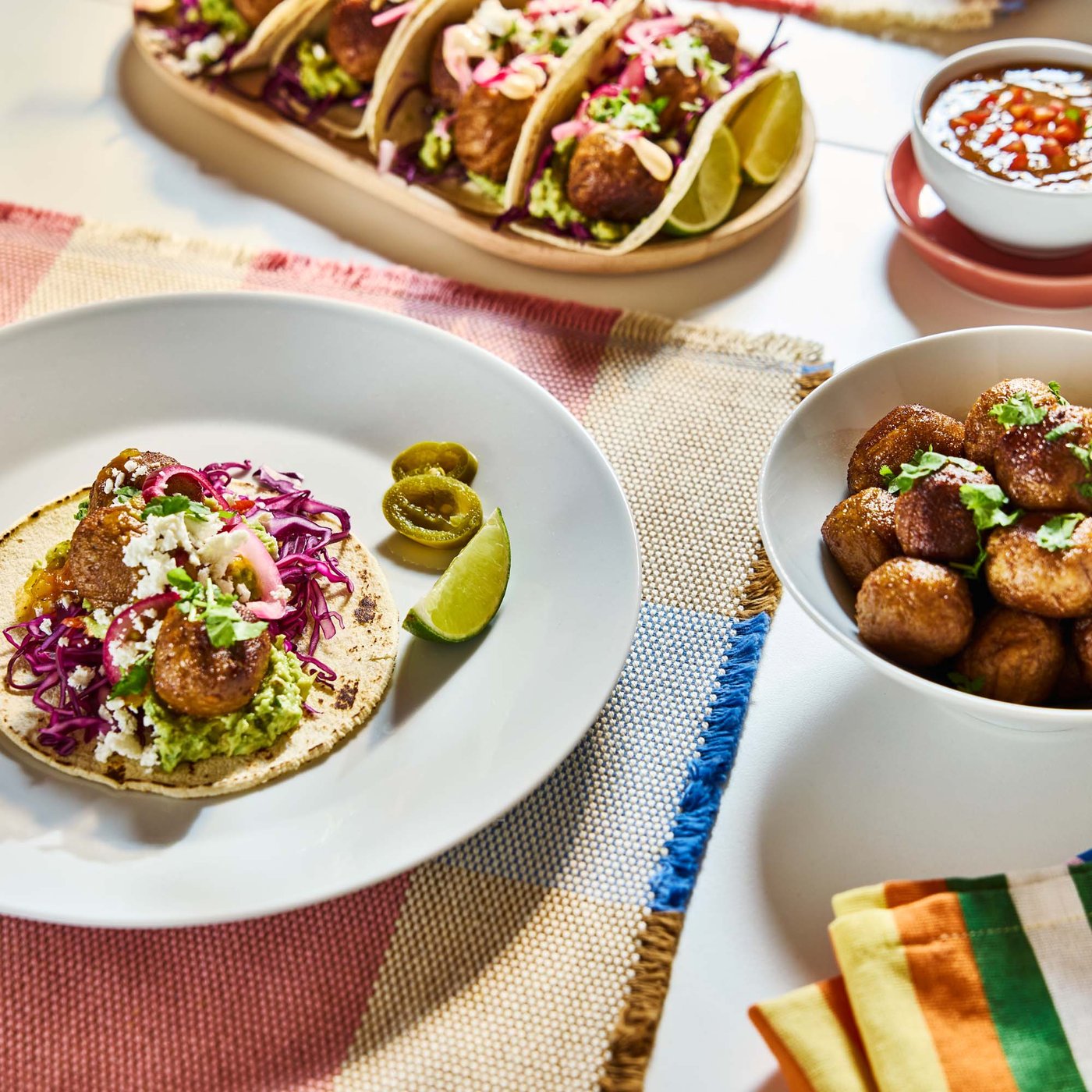 Close-up of a table with bright place mats and a bowl of HUVUDROLL plant balls, next to a plate with plant balls and garnish on a tortilla.