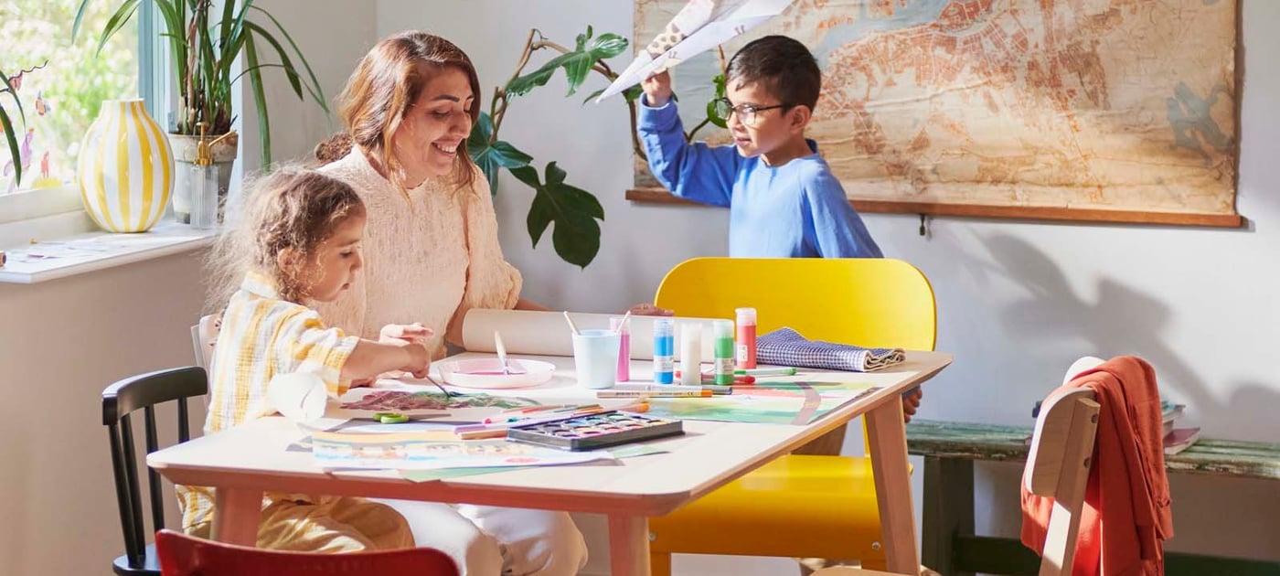 A dining area in a home, where two children and an adult are doing arts and crafts with paints and paper.