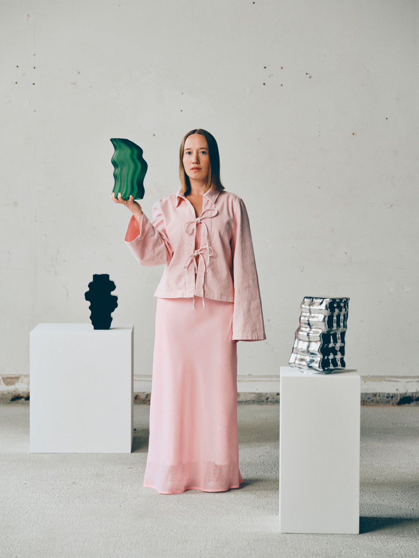 Woman in a pink outfit holding a green abstract sculpture in a minimalist gallery, with additional sculptures displayed on white plinths.