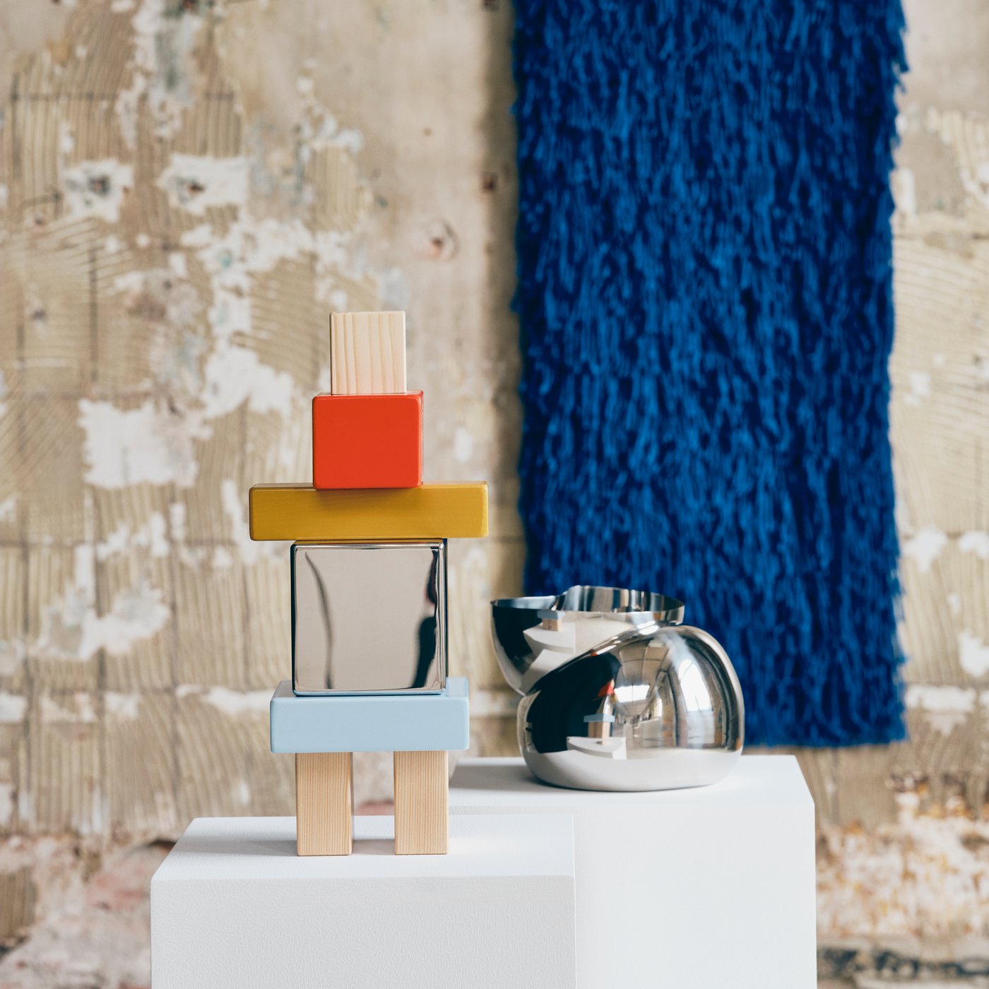 A colourful decorative object and a decorative steel bowl are placed on plinths in an industrial setting with a blue tapestry hanging in the background.