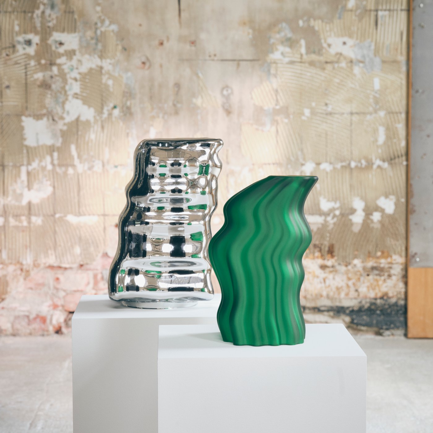 Green and silver abstract sculptures displayed on a white plinth against a textured, weathered wall in an industrial gallery.