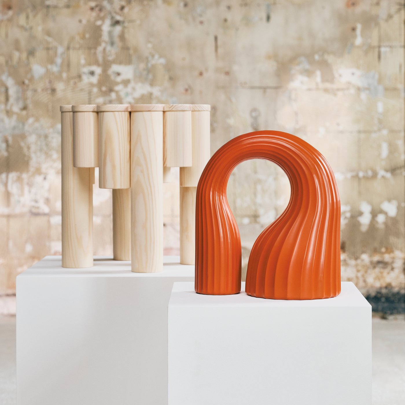 Wooden stool and abstract orange sculpture displayed on white plinths in an industrial gallery.