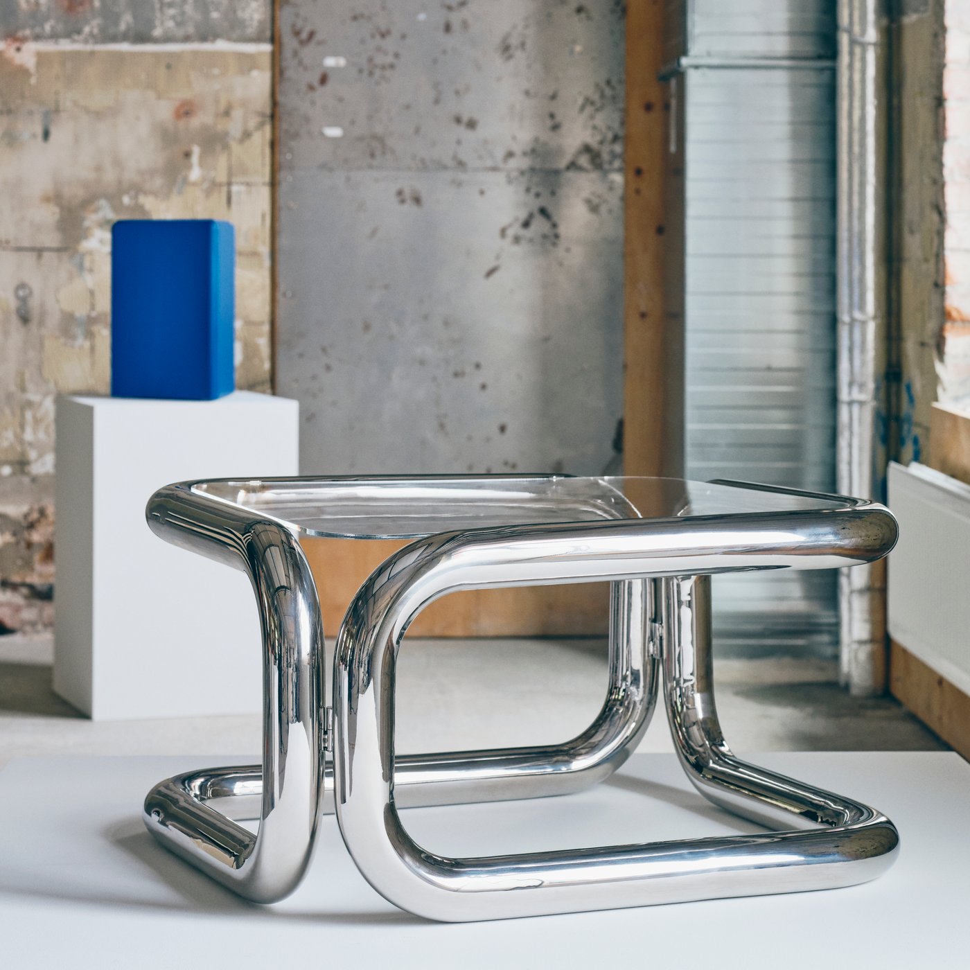 A sculptural chrome table with glass top displayed on a white plinth in an industrial gallery, with a blue geometric sculpture in the background.