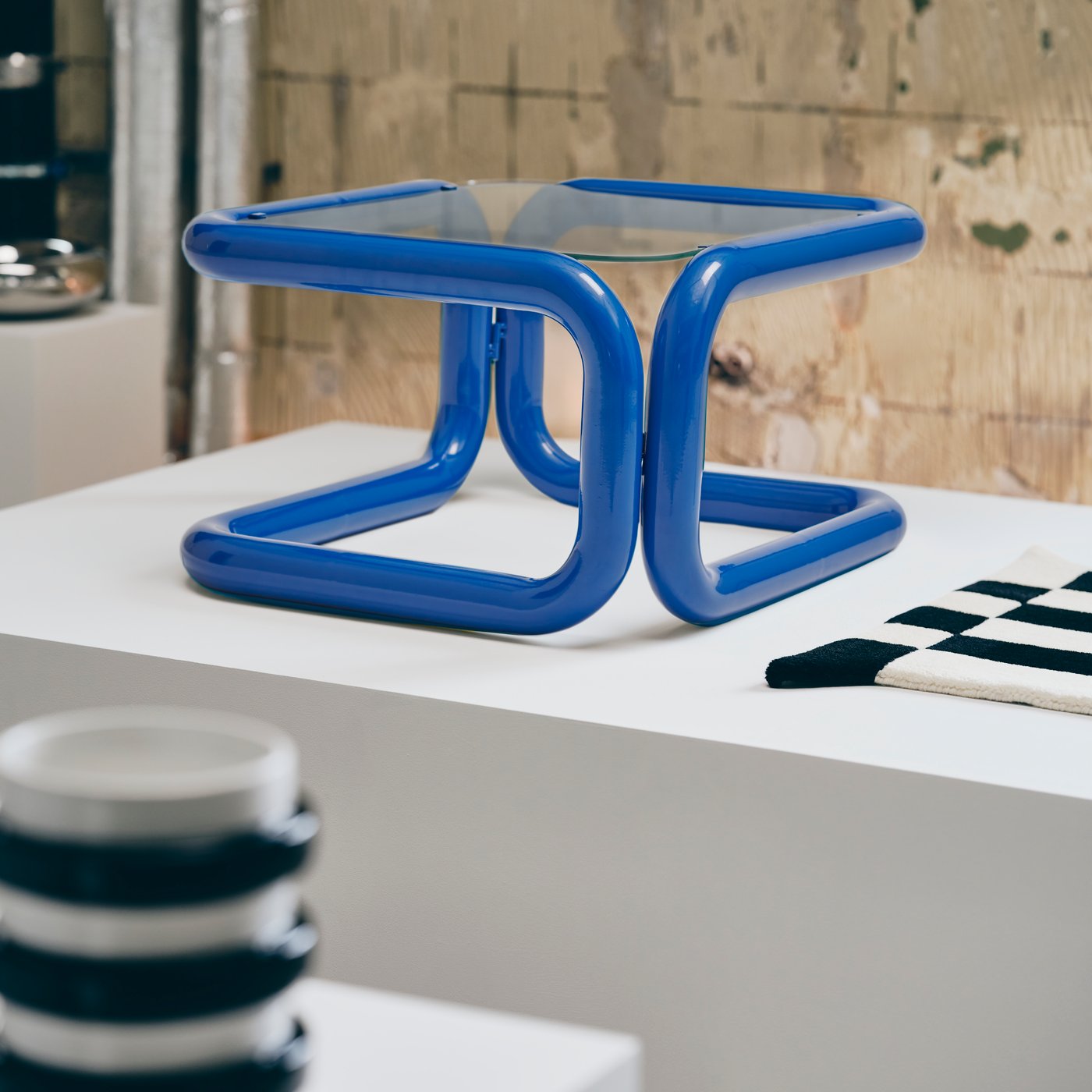 Table with blue base and glass top, displayed on a white plinth in an industrial gallery, with black and white sculpture displayed in the forefront and the edge of a black and white rug in the corner.
